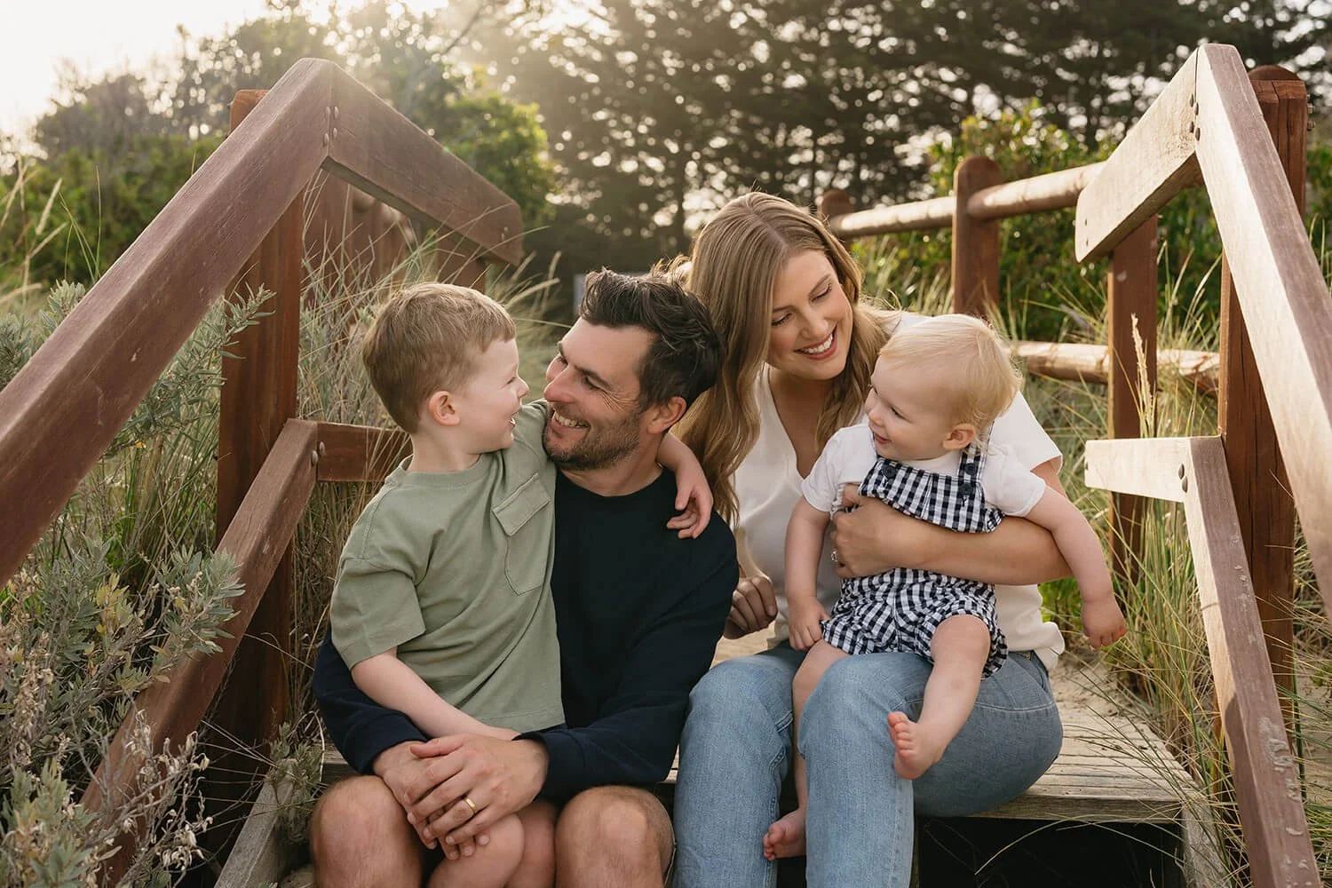 Family of four sitting on wooden steps at Roches Beach Hobart, smiling and interacting in a natural setting with trees and grass.