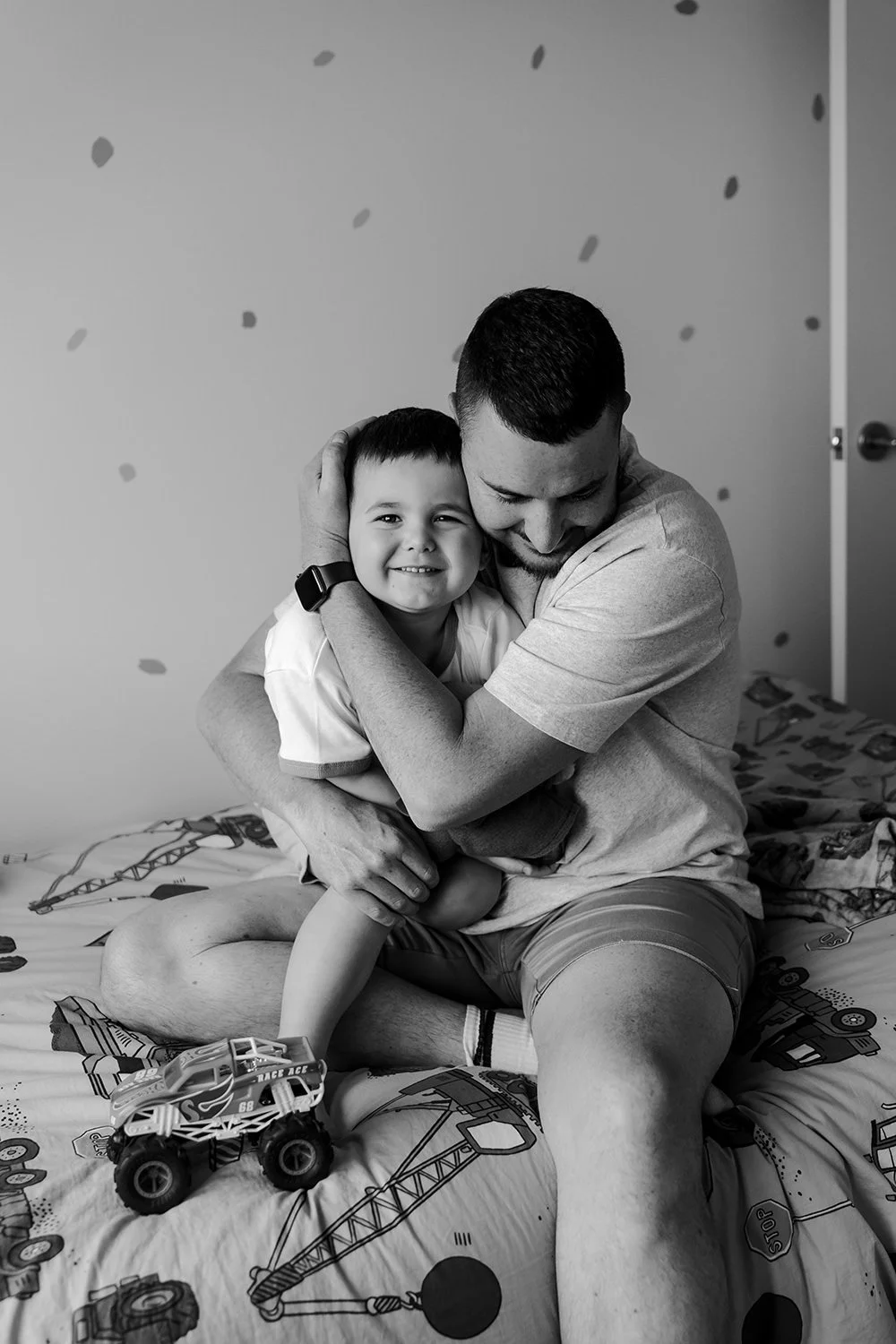 A joyful father and young son hugging in a bedroom with a bed covered in race car-themed bedding and a toy monster truck on the bed.