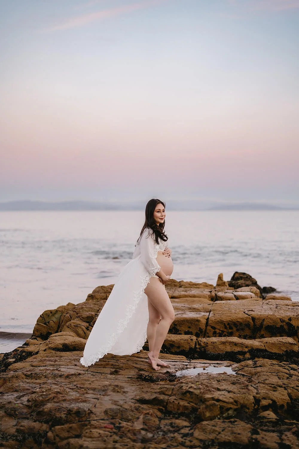 Pregnant woman standing on rocky shore at sunset with ocean in background, wearing a white lace robe and smiling.