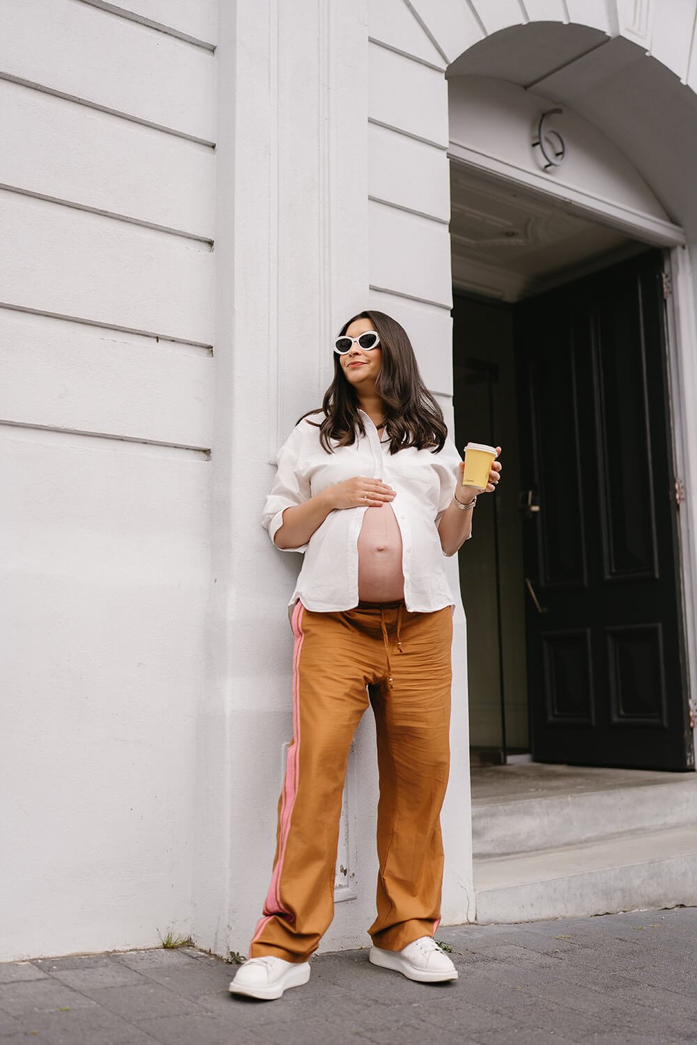 Pregnant woman with long dark hair wearing white sunglasses, white open shirt revealing her pregnant belly, brown pants with pink stripe, holding a yellow coffee cup, standing outside near a white building and black door.