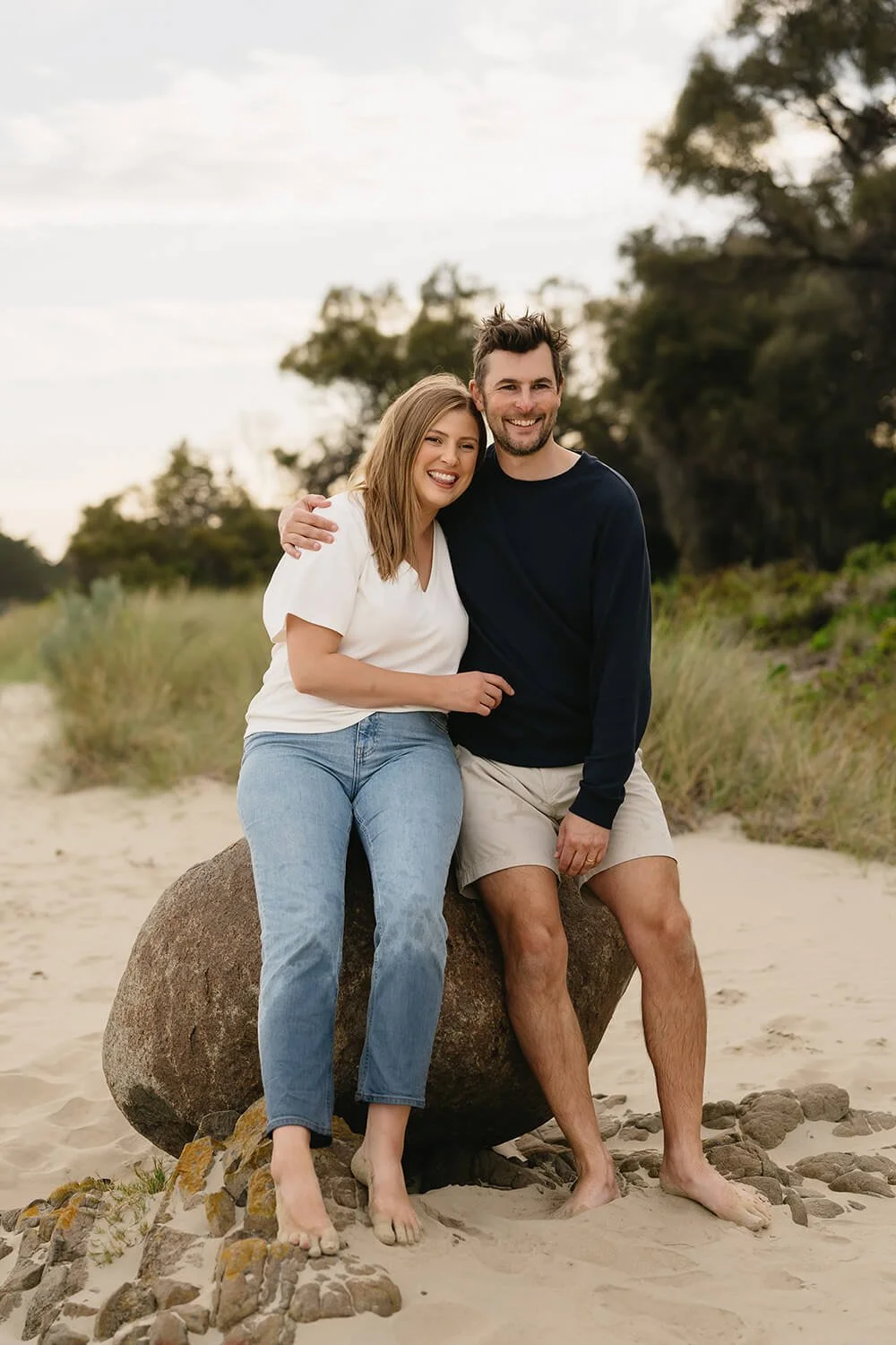 A smiling young couple sitting on a large rock at Roches beach Hobart, with sand, some rocks, grassy plants, and trees in the background.