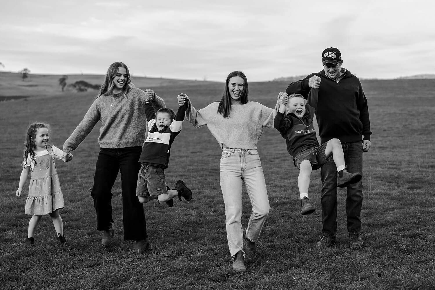Black-and-white photo of a family laughing and playing outdoors in a field. Three adults and three children are holding hands and jumping in the air. The background features rolling hills and a cloudy sky.