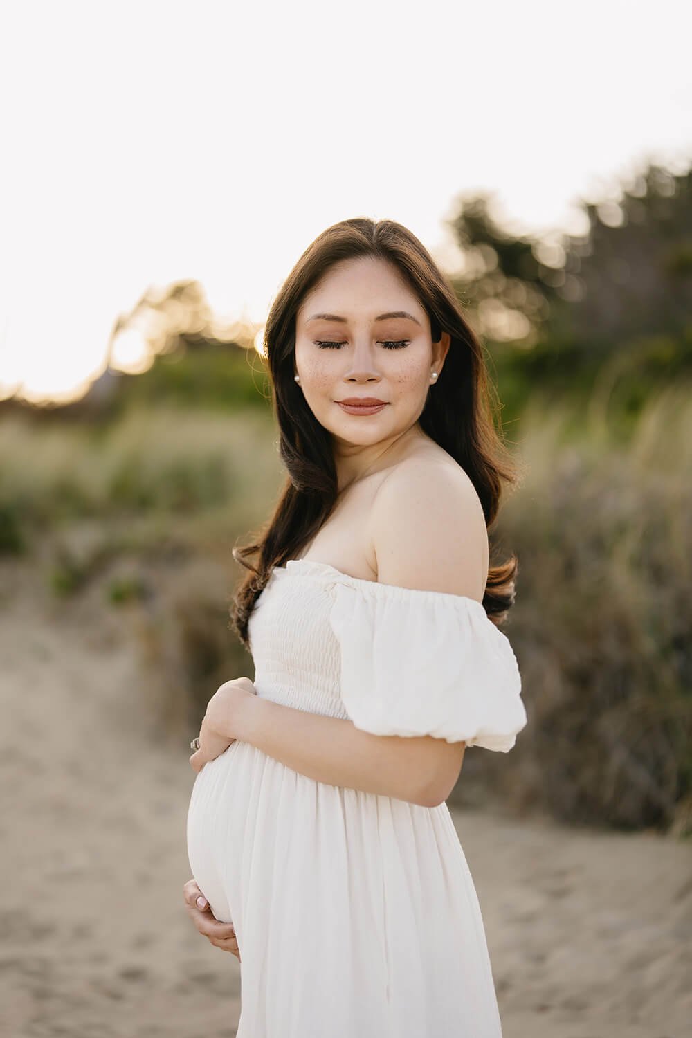 A pregnant woman with long dark hair in a white off-the-shoulder dress standing outdoors on a trail, with greenery and sun setting in the background, eyes closed and smiling softly.