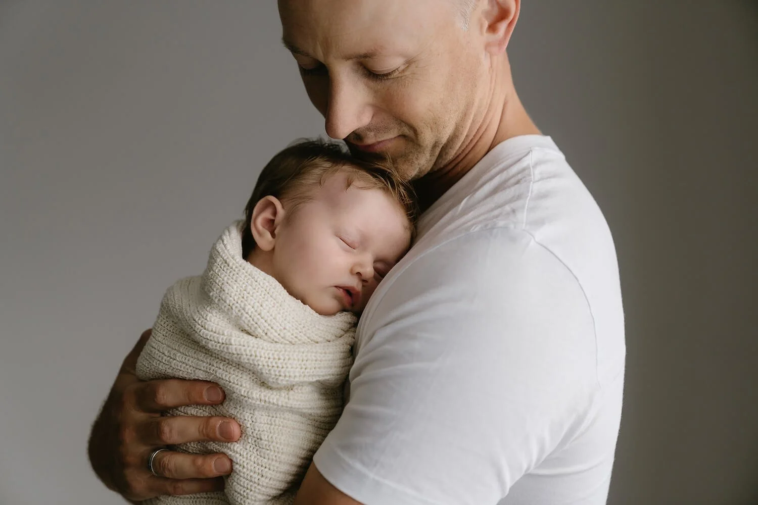 A man gently holding a sleeping baby wrapped in a beige blanket, with a neutral gray background.