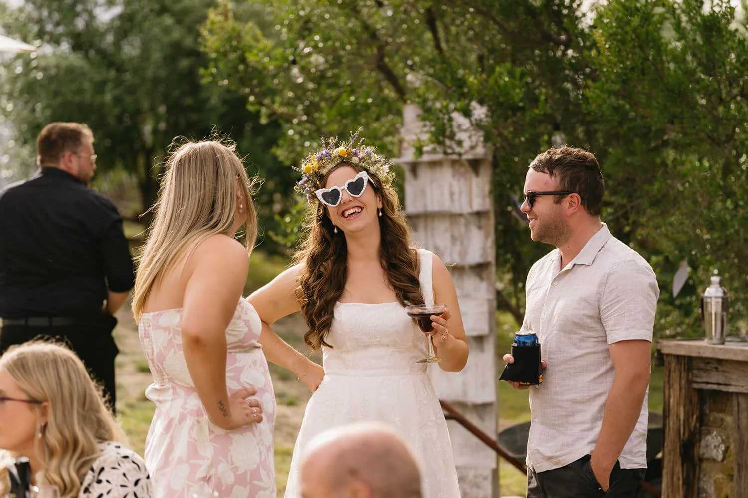 People socializing outdoors at a wedding reception, woman in floral crown and sunglasses holding a drink, surrounded by friends. One woman in pink dress is talking to her, man in light shirt and sunglasses is smiling.
