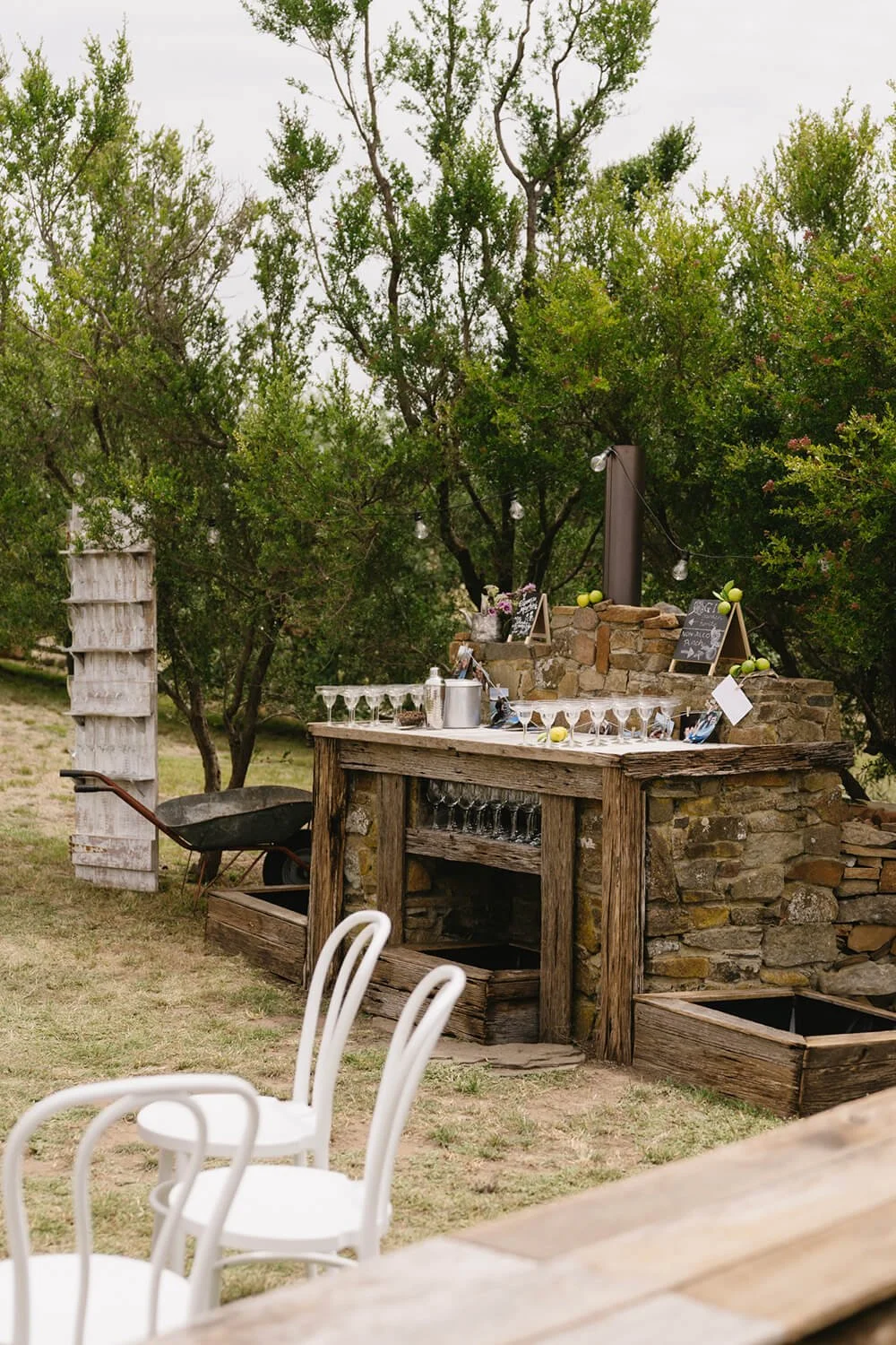 Outdoor rustic bar setup with wine glasses, chalkboard signs, and string lights amid trees.