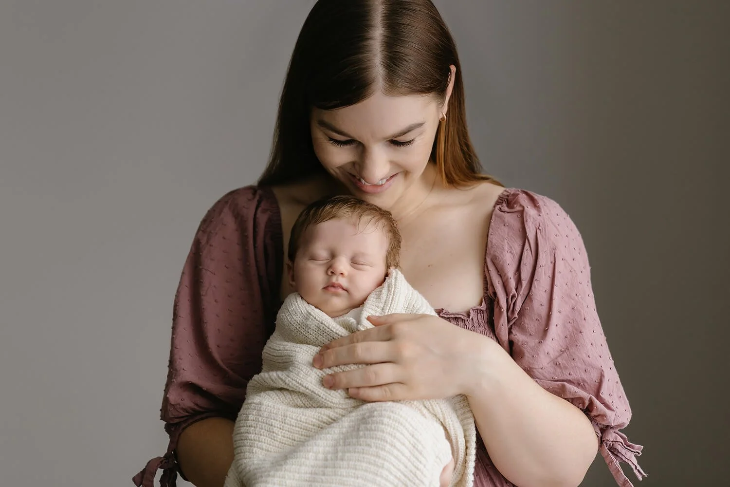 A woman with brown hair holding a sleeping baby wrapped in a cream blanket, both smiling gently, against a plain grey background.