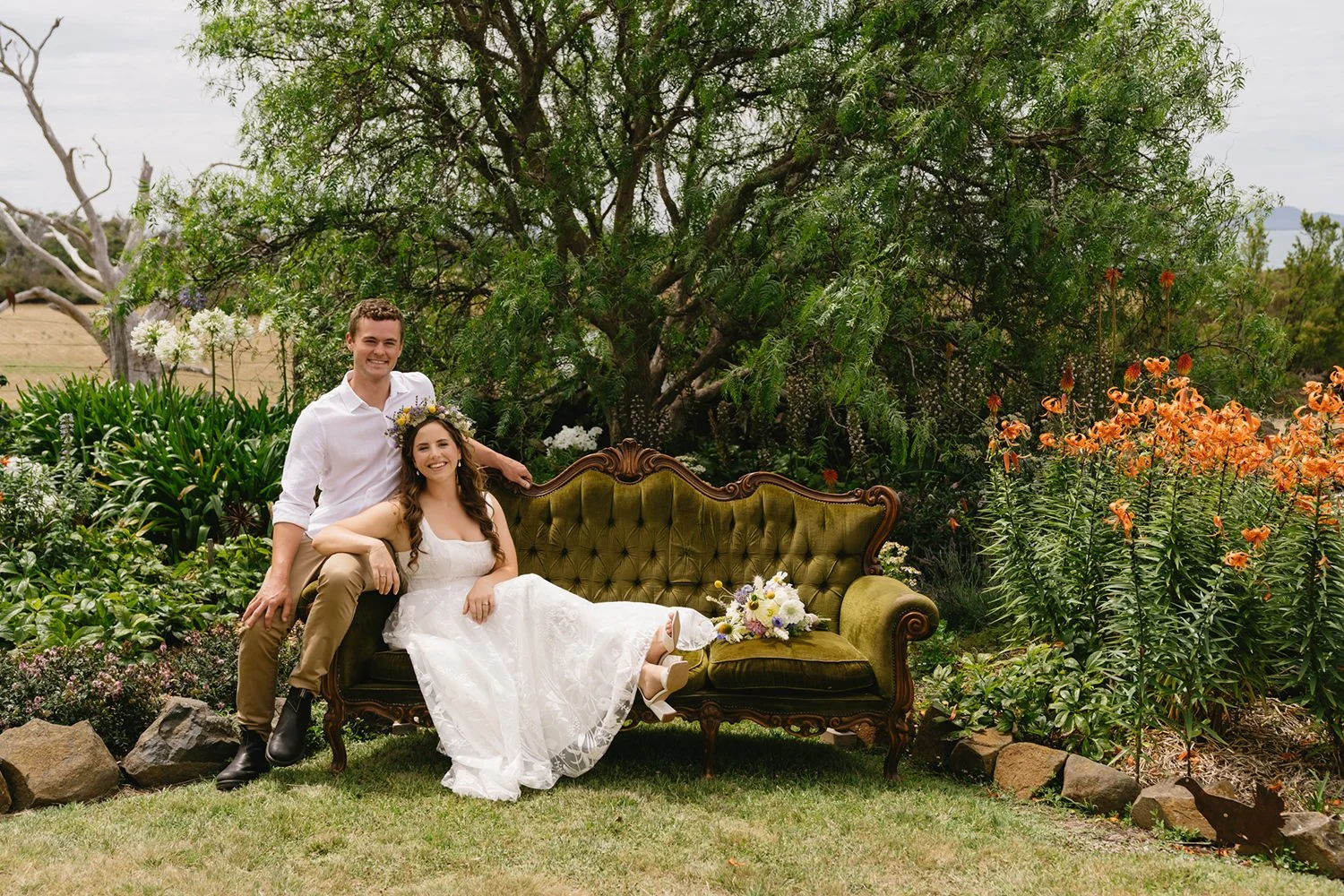 A smiling bride and groom sitting on a vintage green velvet sofa outdoors surrounded by greenery and orange flowers. The bride is wearing a white lace wedding dress with a floral crown, and the groom is in a white shirt and khaki pants. There is a bo