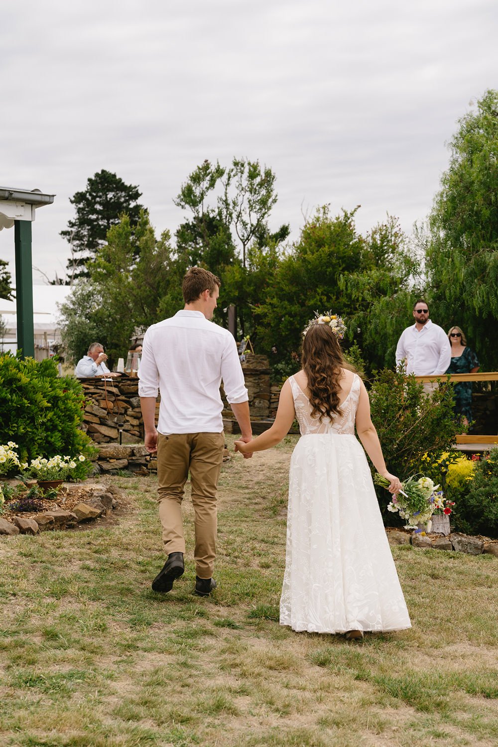A couple holding hands walking outdoors at a wedding ceremony, with guests and lush greenery in the background.