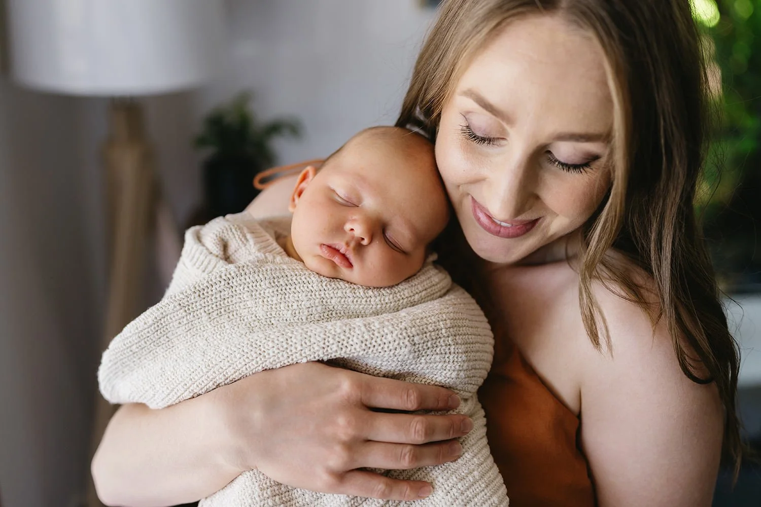 A woman holding a sleeping newborn baby wrapped in a beige knitted blanket, both displaying affection and contentment.
