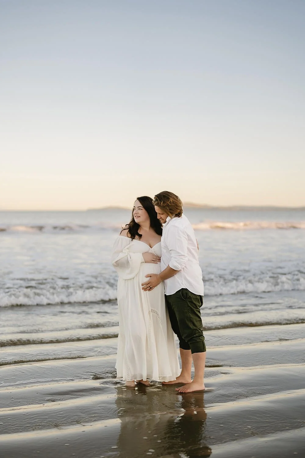 A couple standing on the beach at sunset, with the woman wearing a white dress and the man in a white shirt and dark pants, holding hands and touching stomachs.