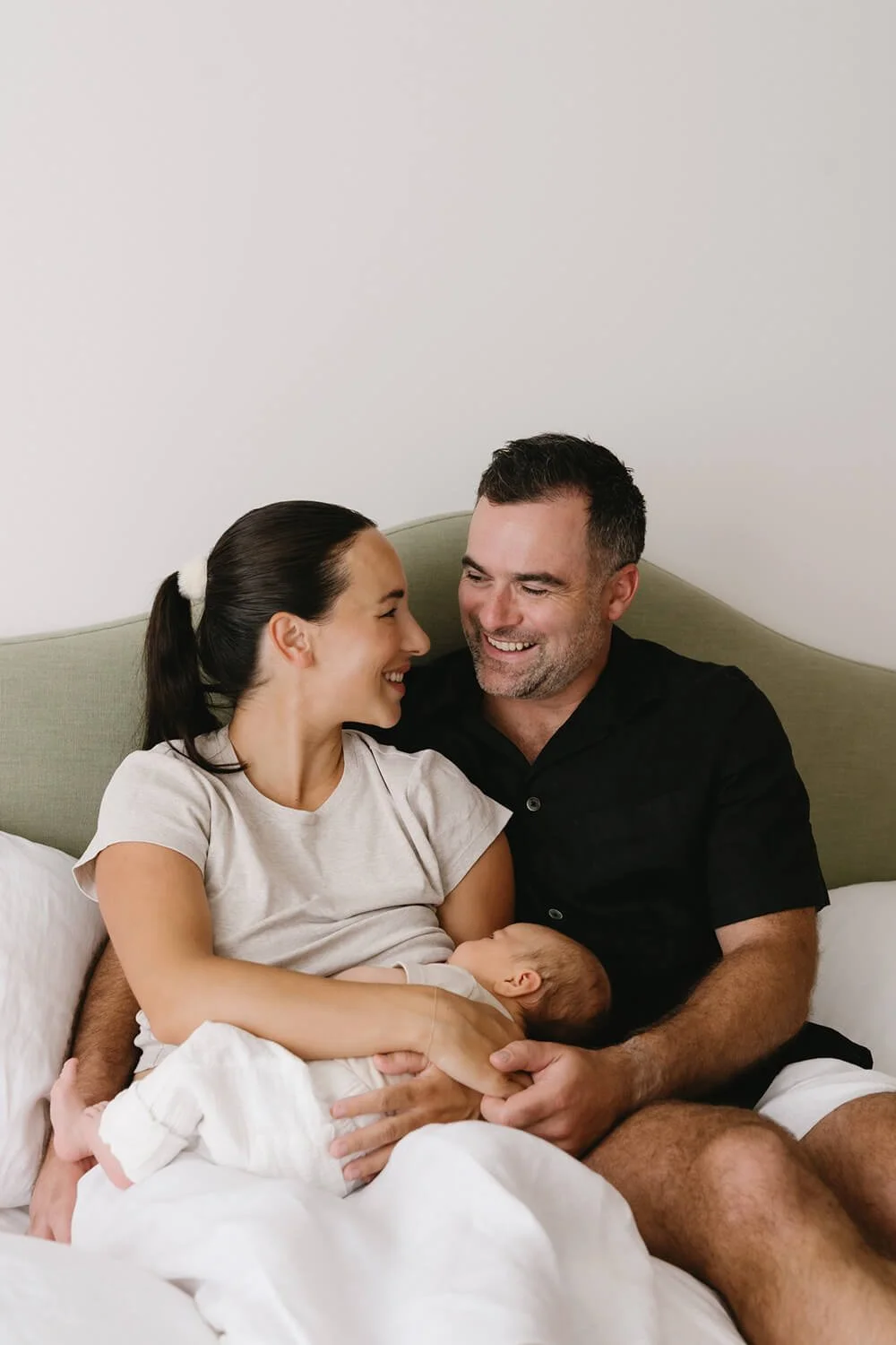 A happy family of three, a man, a woman, and a baby, sitting on a bed in a cozy room, sharing a joyful moment together.