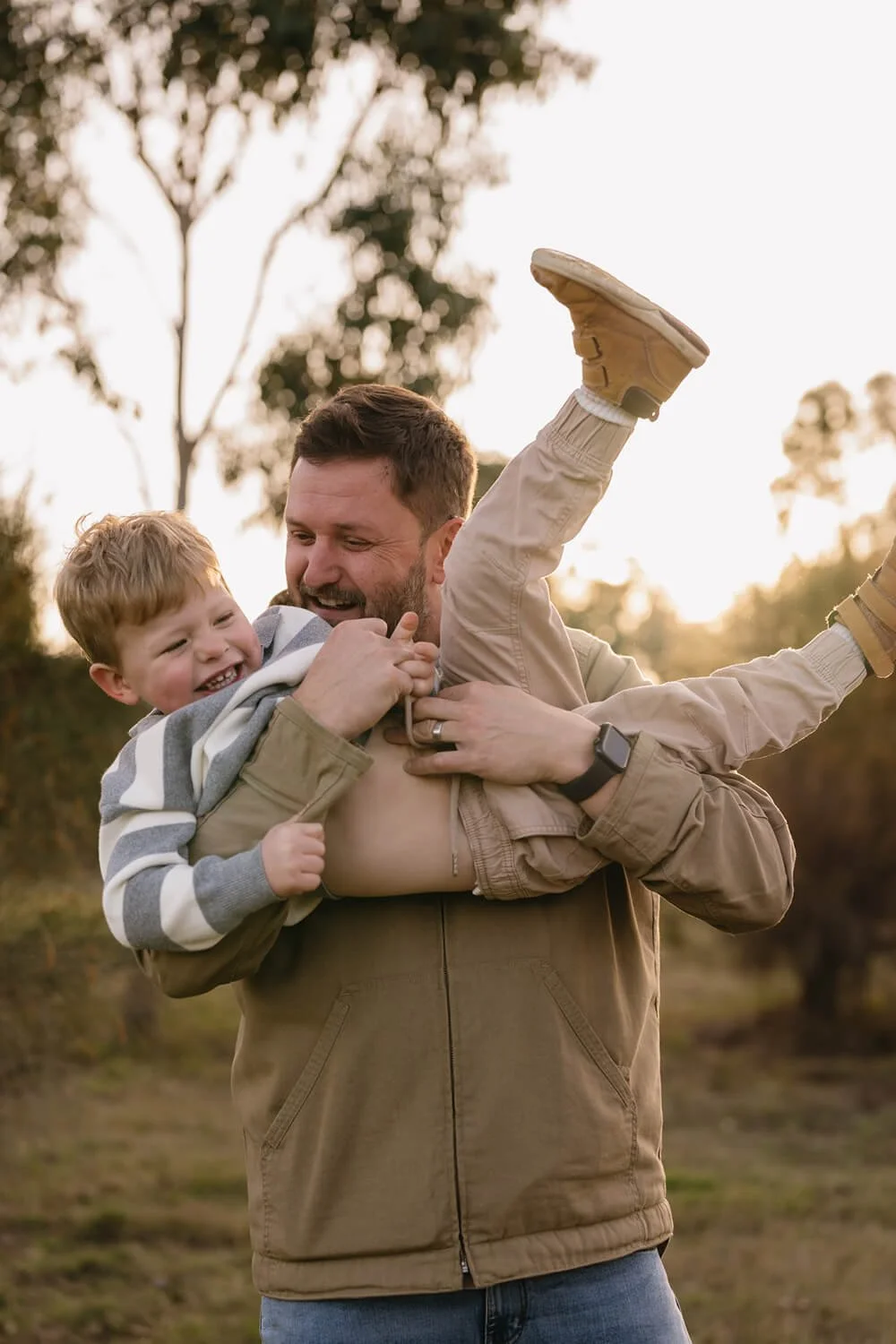 A man with a beard wearing a brown jacket is playfully carrying a young boy with red hair, who is smiling and laughing, outdoors during sunset. The boy's legs are thrown in the air as they enjoy a playful moment.