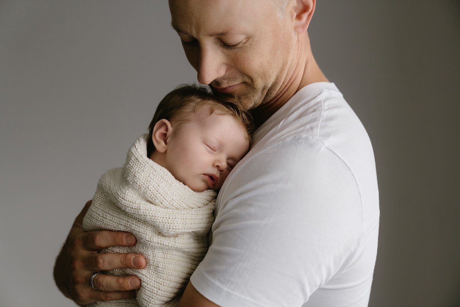 A father holding a sleeping baby wrapped in a knitted blanket, with the father's head gently resting against the baby's head, against a plain neutral background.