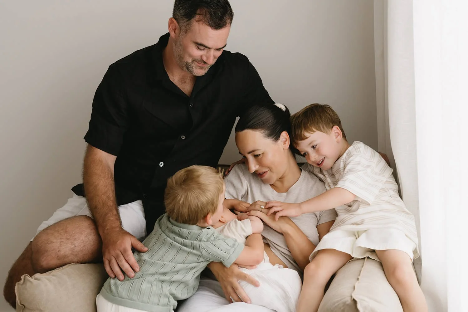 A family of five gathered around a woman sitting on a couch, with children touching her and a man leaning over her, all smiling and enjoying a joyful moment together.