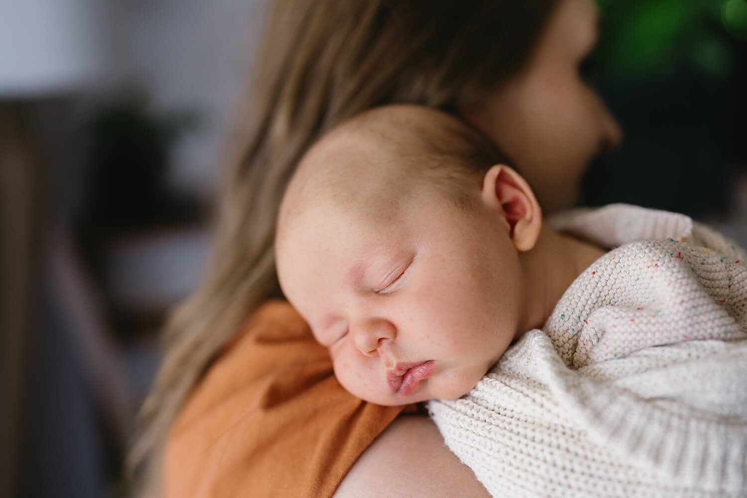 A young child is peacefully sleeping on a woman's shoulder, with her face slightly out of focus in the background.