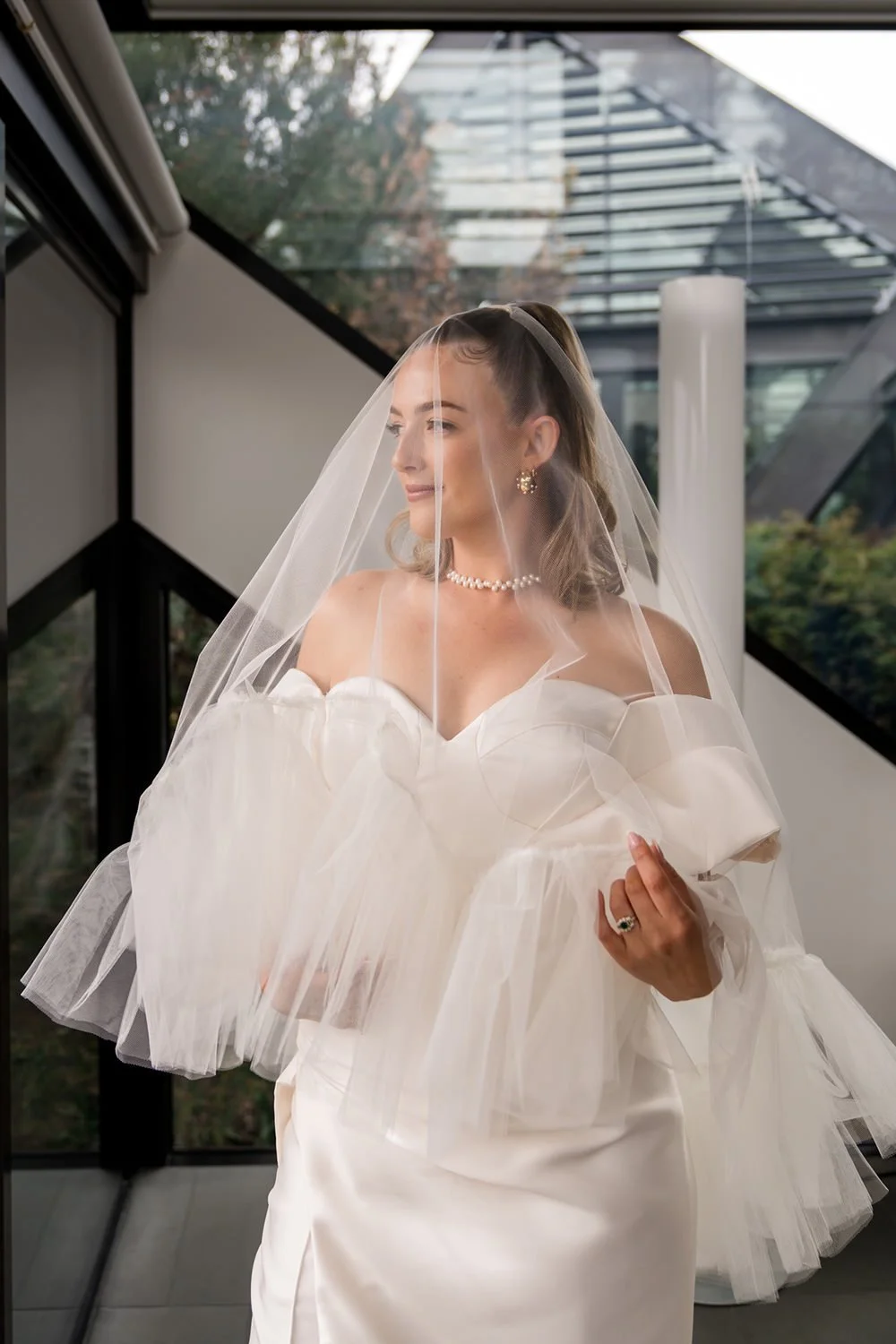 A bride in a white wedding dress with puffy sleeves and a veil, standing indoors with a modern glass building in the background.