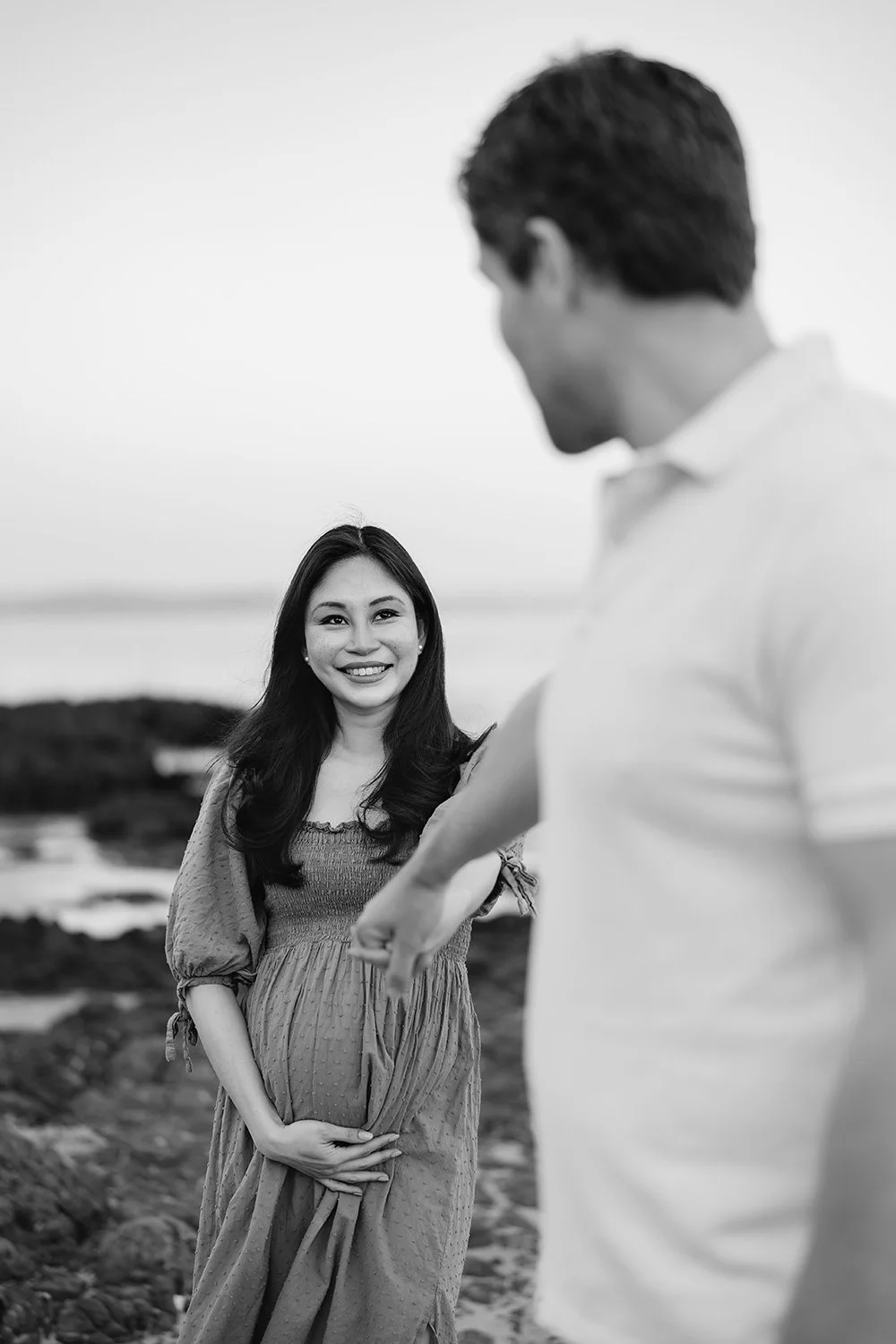 A smiling pregnant woman with long dark hair, wearing a dress, standing outdoors by water. A man, slightly out of focus, is gesturing towards her with his arm extended.