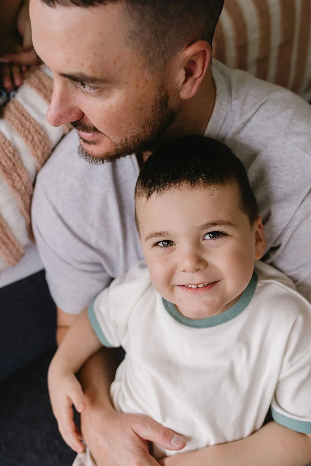 A father and son sitting close together. The father has a beard and a haircut, wearing a light gray shirt, while the son has short dark hair, a smile, and is wearing a white t-shirt with light blue cuffs and collar.