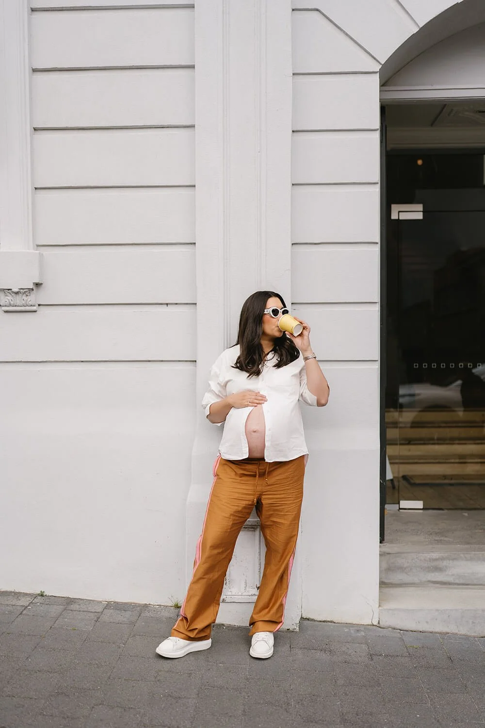 Pregnant woman wearing white blouse, brown pants, white sneakers, and sunglasses, drinking from a cup while standing against a white wall outside a building.