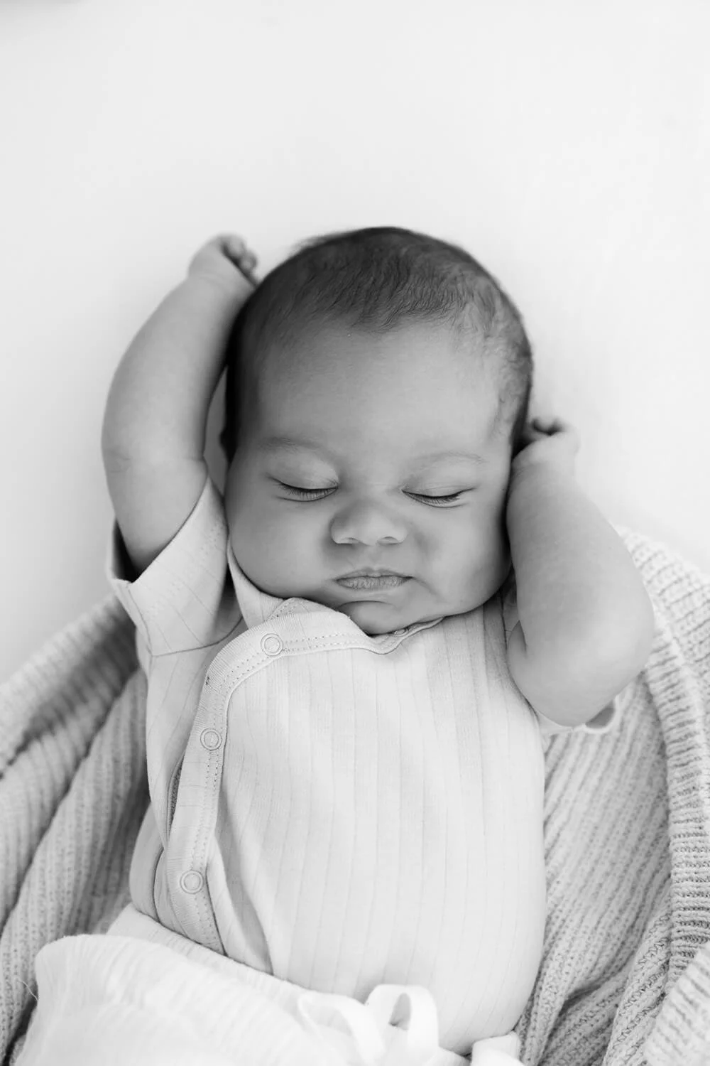 A sleeping baby with arms raised behind the head, resting on a blanket, in black and white.