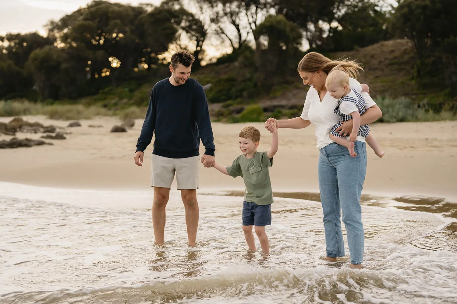 A family enjoying a walk on Roches beach Hobart, holding hands in the shallow water, with trees and a hill in the background.