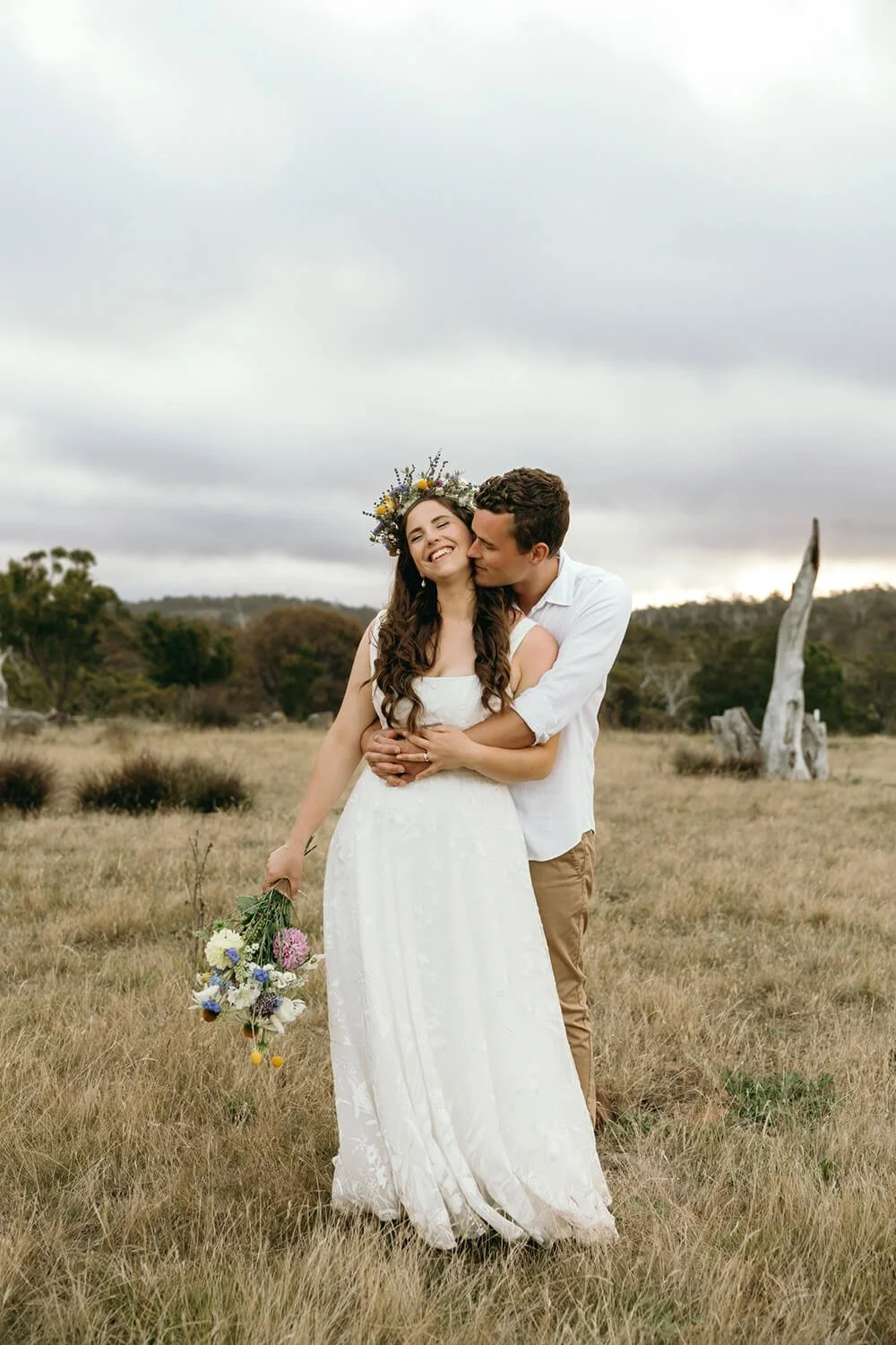 A couple in wedding attire embraces in a grassy field under a cloudy sky, with the woman wearing a white dress and a floral crown, holding a bouquet, and the man in a white shirt and tan pants, kissing her cheek.