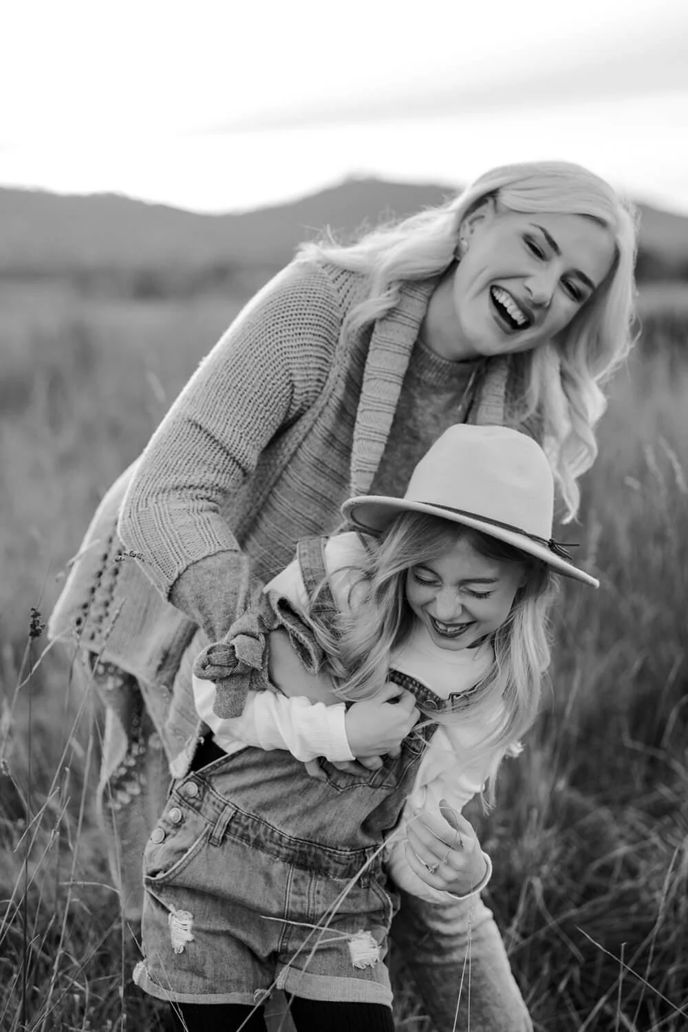 A woman and a young girl playing and laughing together in a grassy field, with mountains in the background, black and white photo.