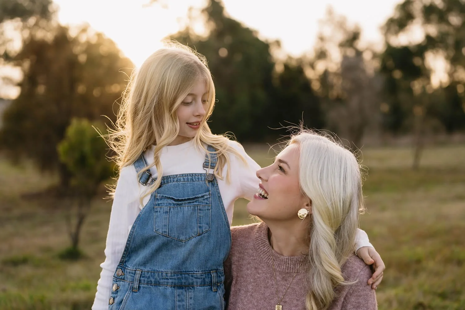 A woman with blonde hair and earrings kneels in a park at sunset, looking up and smiling at a young girl with long blonde hair who stands next to her, wearing denim overalls.