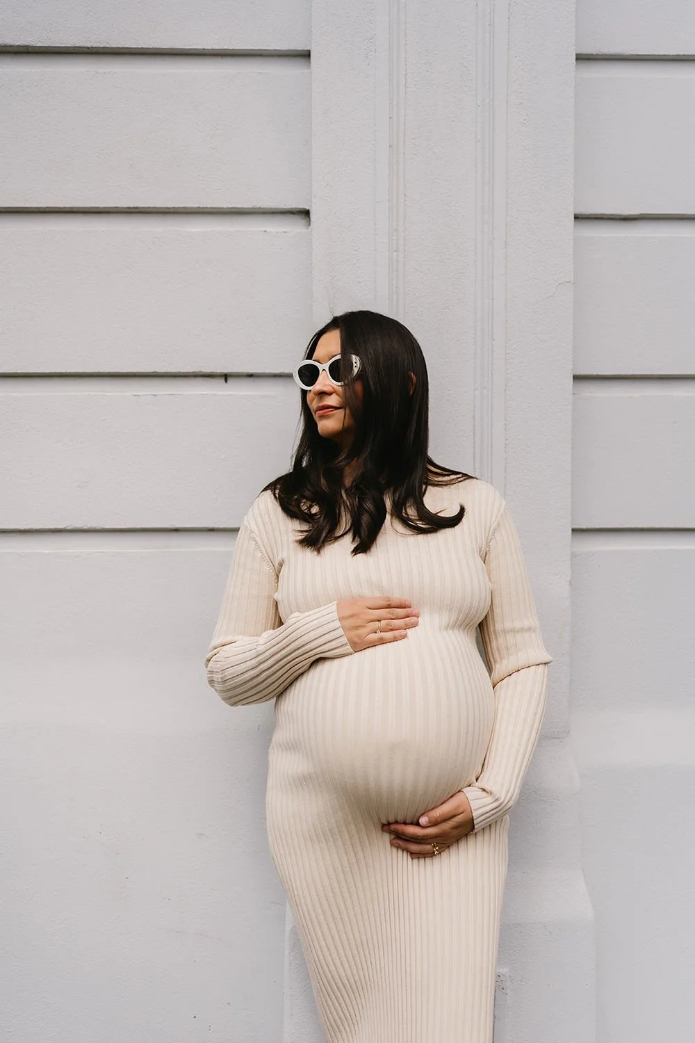 A pregnant woman wearing a beige, ribbed dress and white sunglasses standing against a white textured wall, gently holding her belly with her right hand and smiling softly.