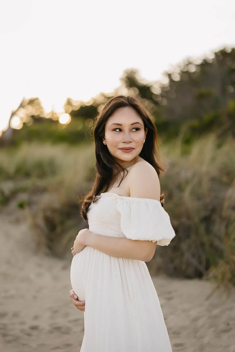 Maternity photo of a woman with dark hair, wearing a white off-shoulder dress, standing on a sandy beach at sunset with trees in the background.