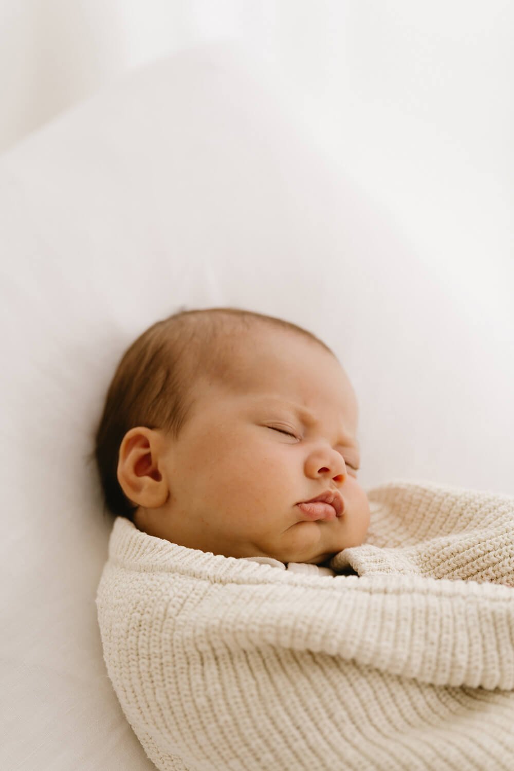 A sleeping baby wrapped in a cream knitted blanket, resting on a white surface.