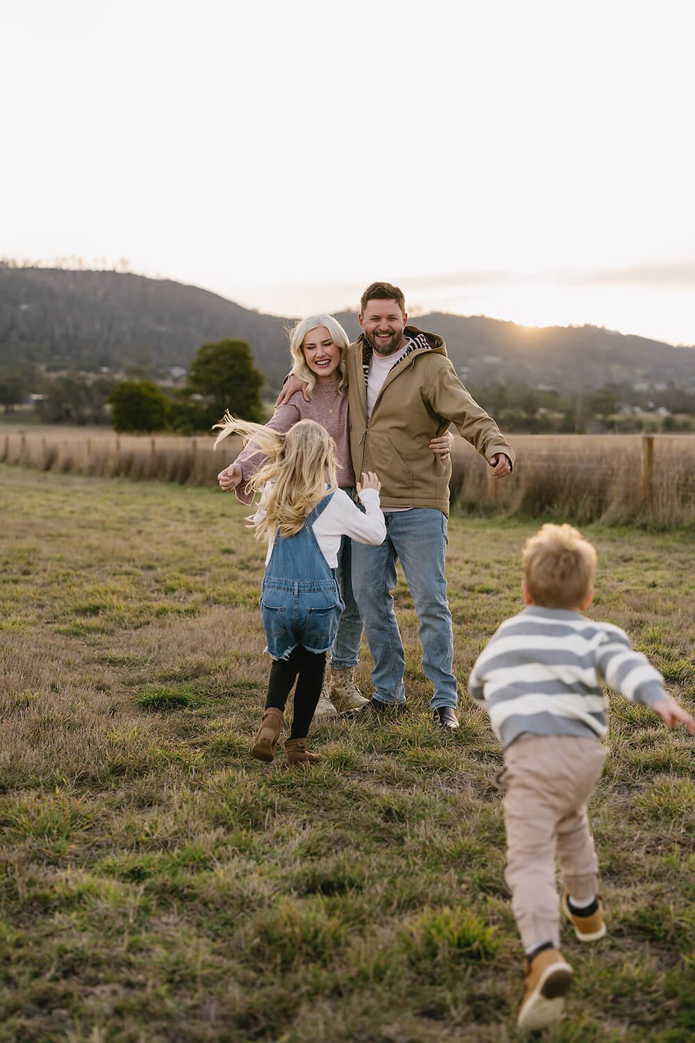 A joyful family of four playing in a grassy field during sunset, with mountains in the background.