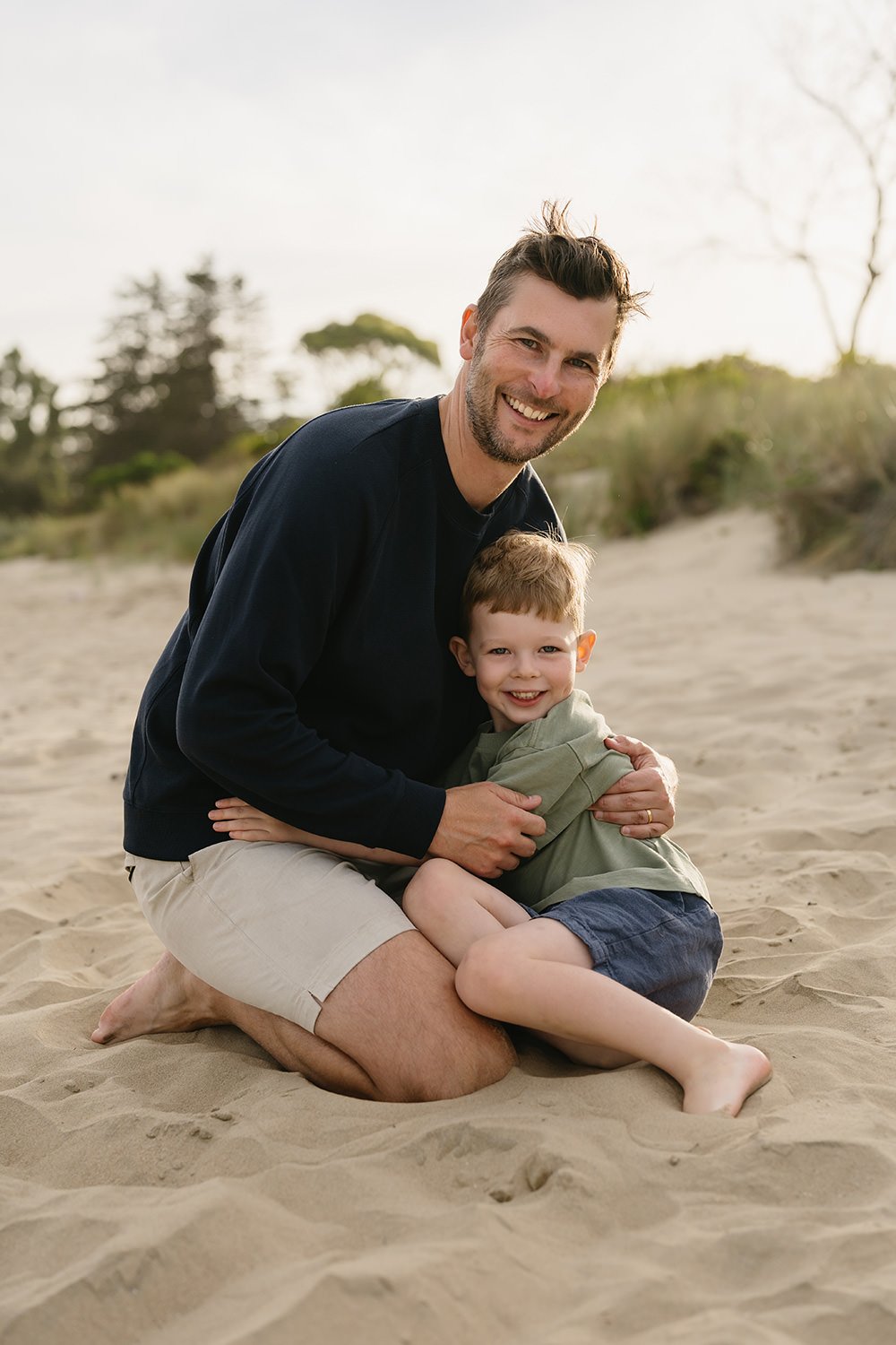A happy man and boy sitting on the sand at the beach, smiling at the camera. The man is wearing a black long-sleeve shirt and beige shorts, and the boy is wearing a green hoodie and dark shorts.