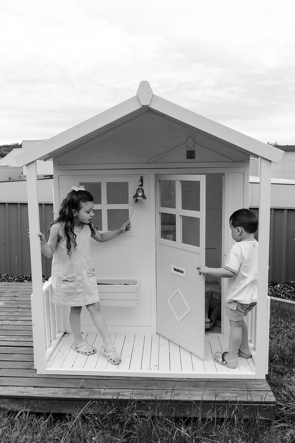 Two children playing in a small outdoor playhouse, with one girl holding a bell and a boy opening the door, on a wooden deck.