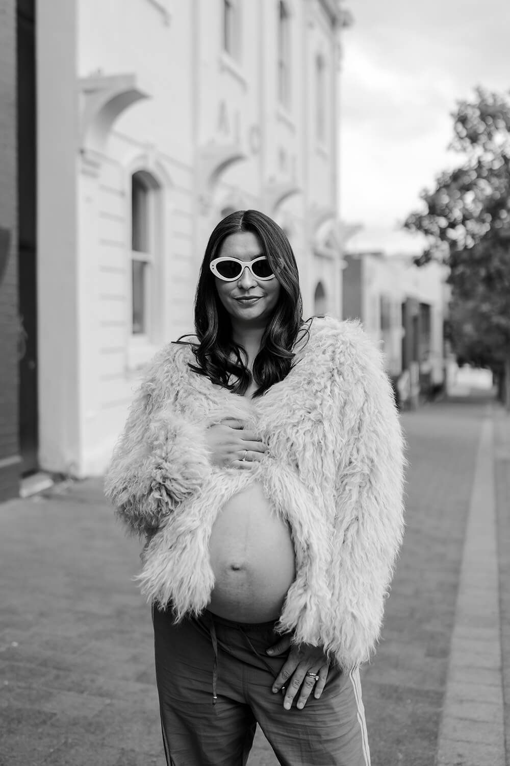 A woman standing outdoors on a city street, wearing sunglasses, a fluffy fur coat, and sports pants, with a pregnant belly visible.