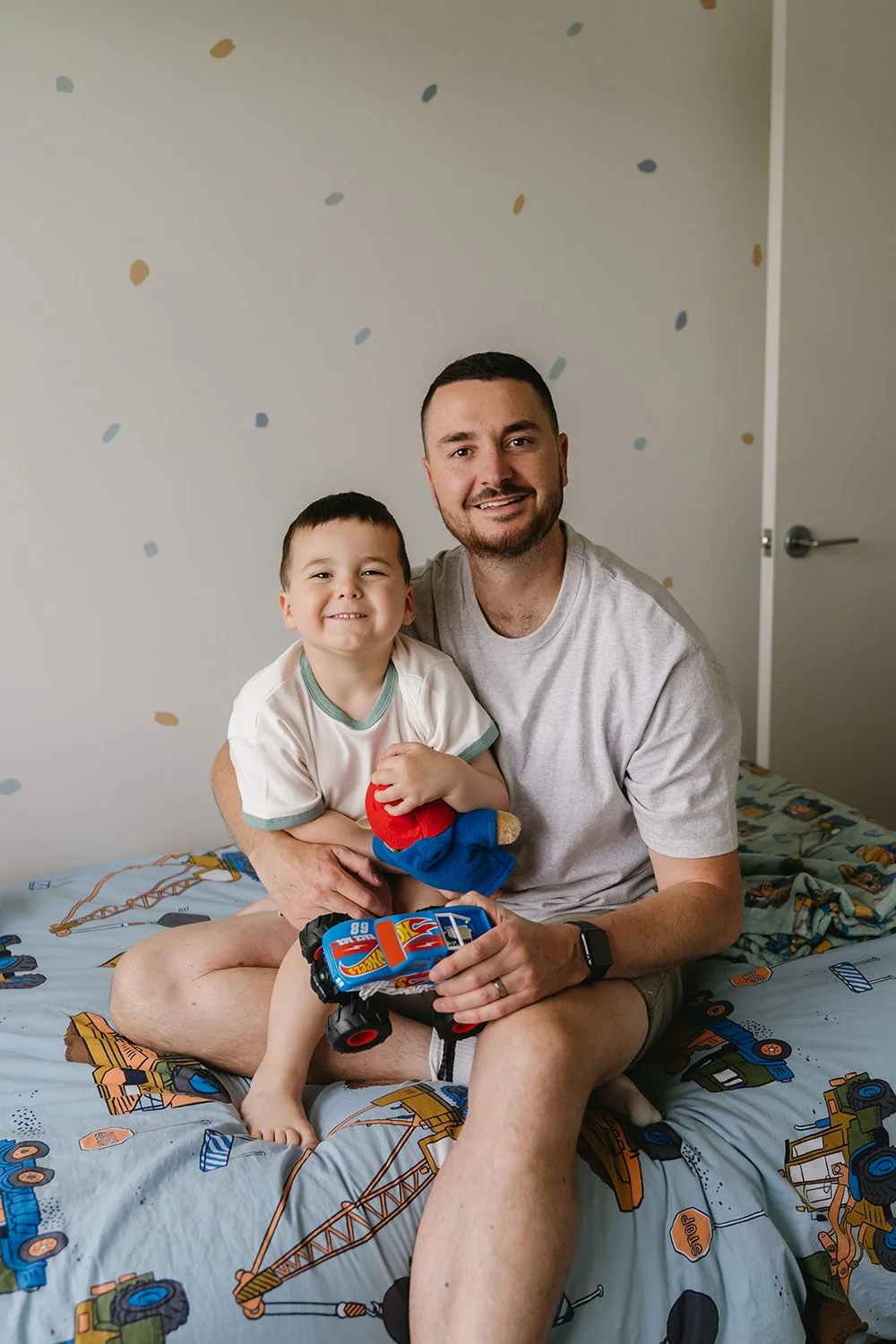 A man and a young boy sitting on a bed with construction-themed bedding. The boy is holding a stuffed toy and a toy car, both smiling at the camera in a warmly lit room with a plain wall decorated with colorful dots.