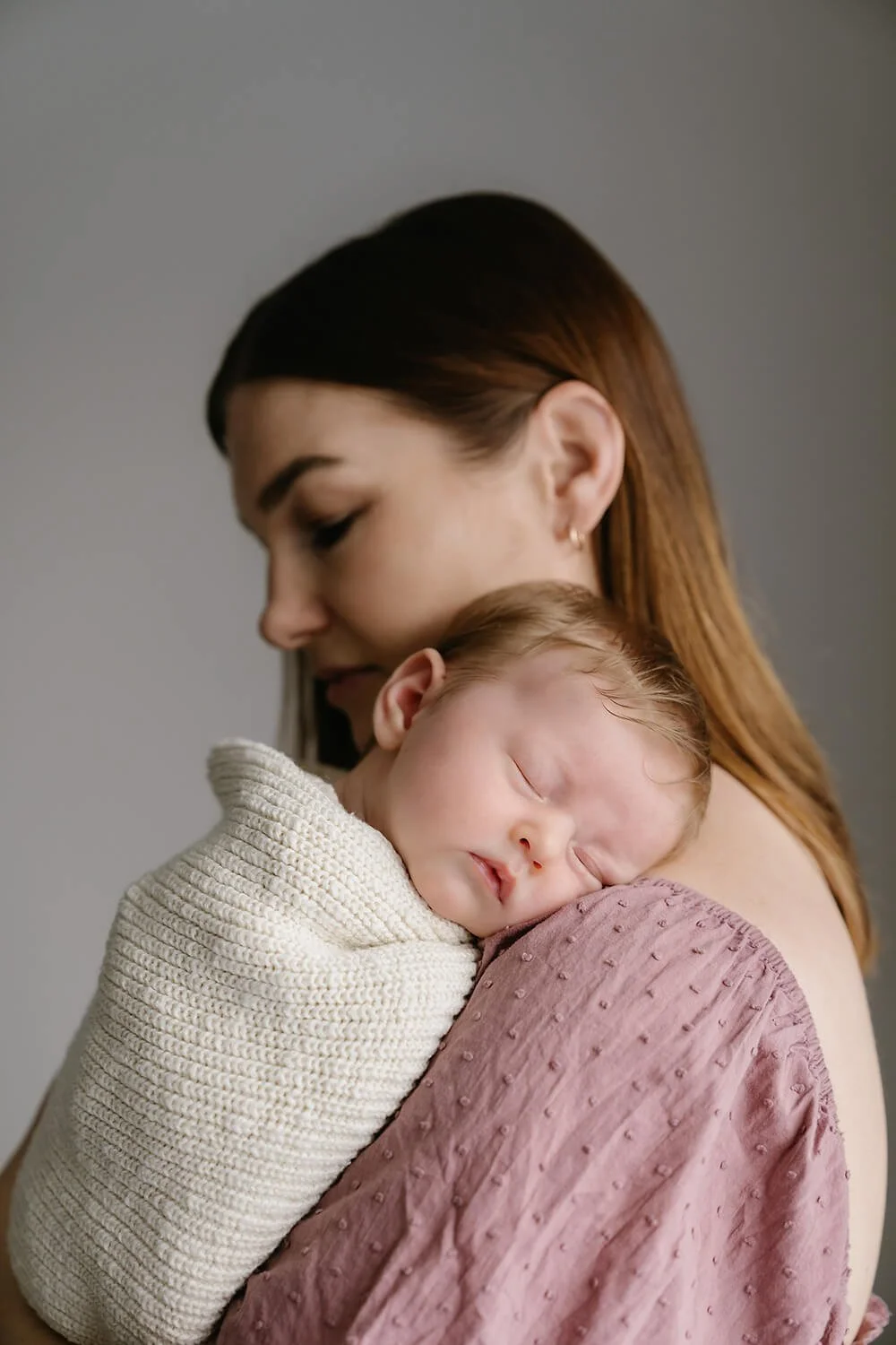 A woman with brown hair holding a newborn baby wrapped in a cream knitted blanket and a mauve textured cloth, both with serene expressions against a plain gray background.