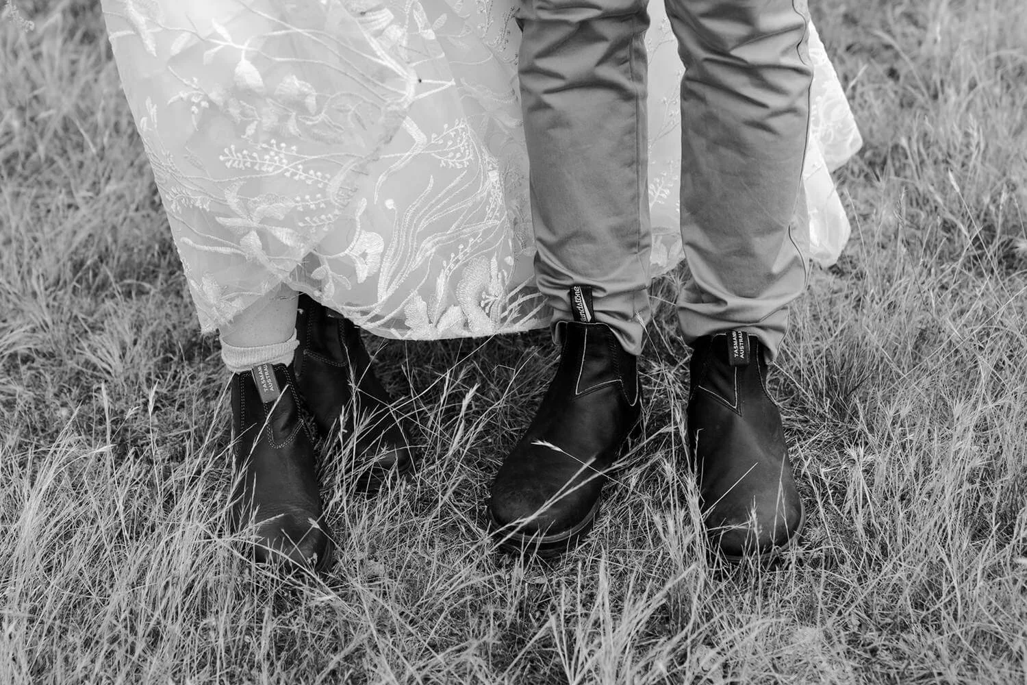Close-up of a couple's lower bodies standing on grass, showing their wedding attire and black boots.