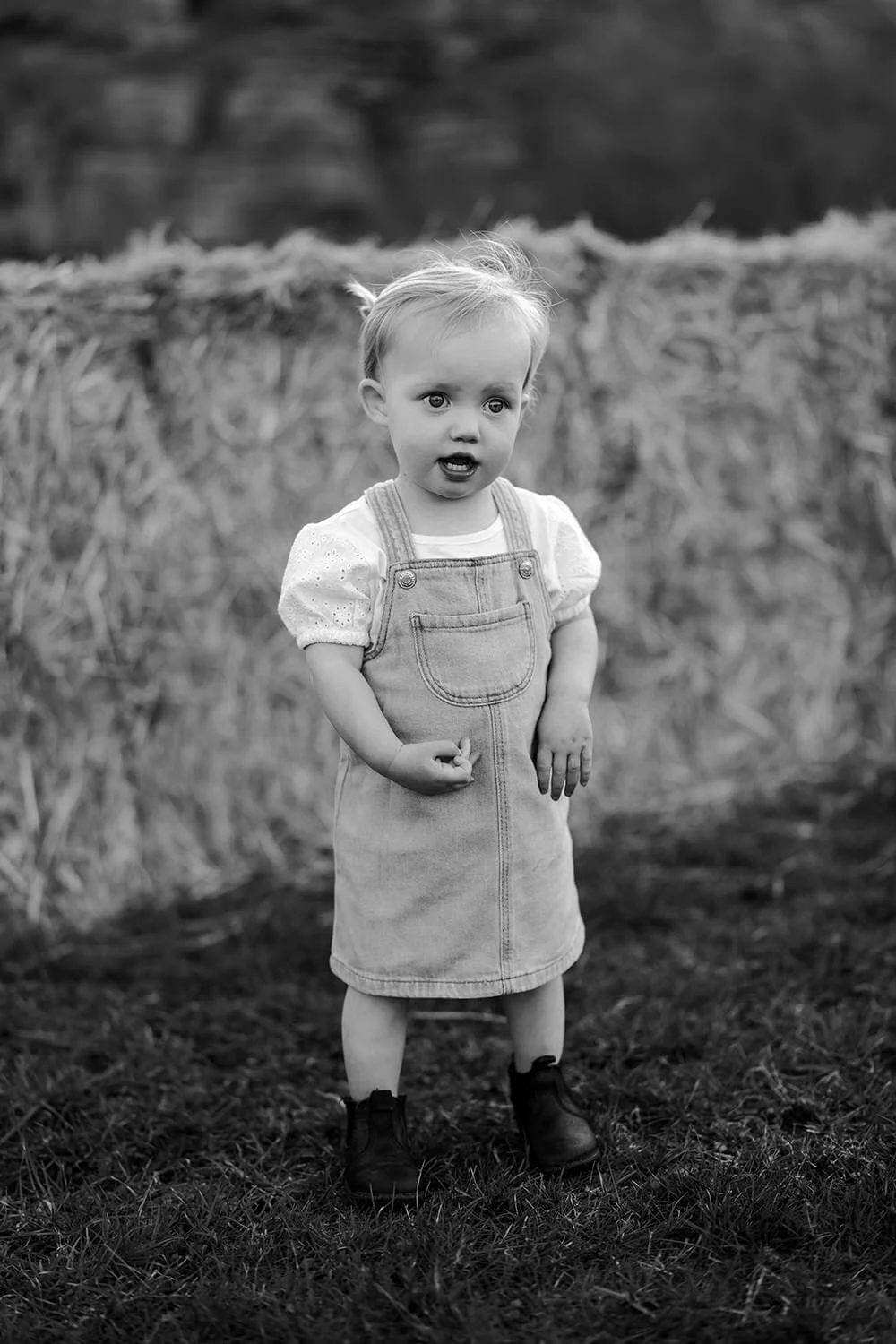 A young girl with light hair, wearing a denim dress over a blouse, standing outdoors on grass with a pile of hay in the background.