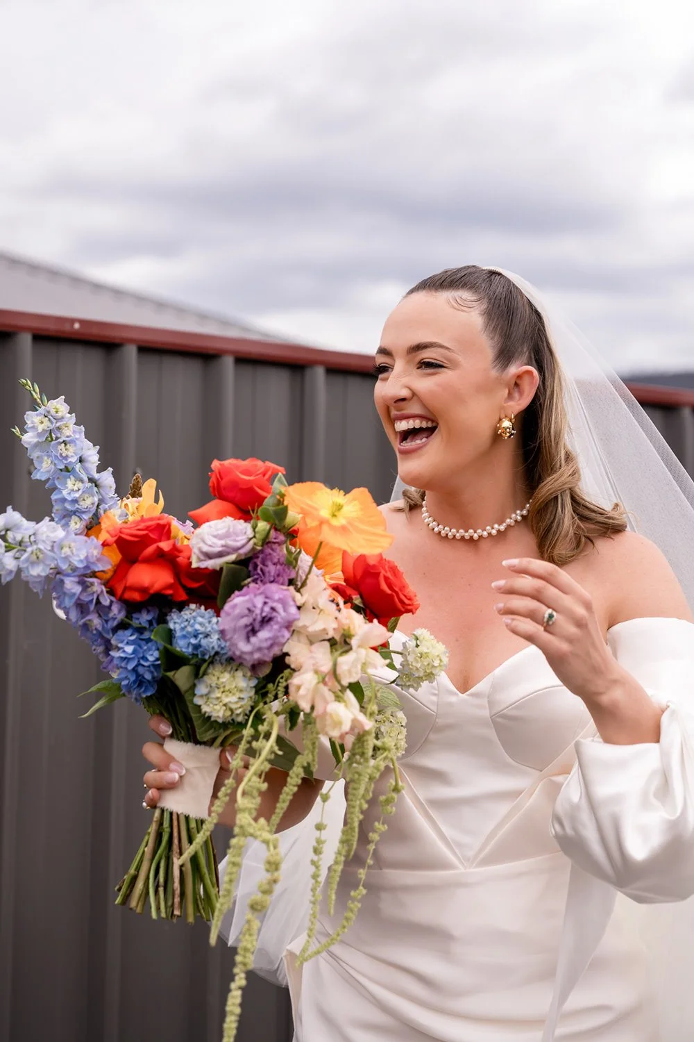 A smiling bride in a white off-shoulder wedding dress holding a colorful bouquet of flowers, standing outdoors against a gray fence and cloudy sky.