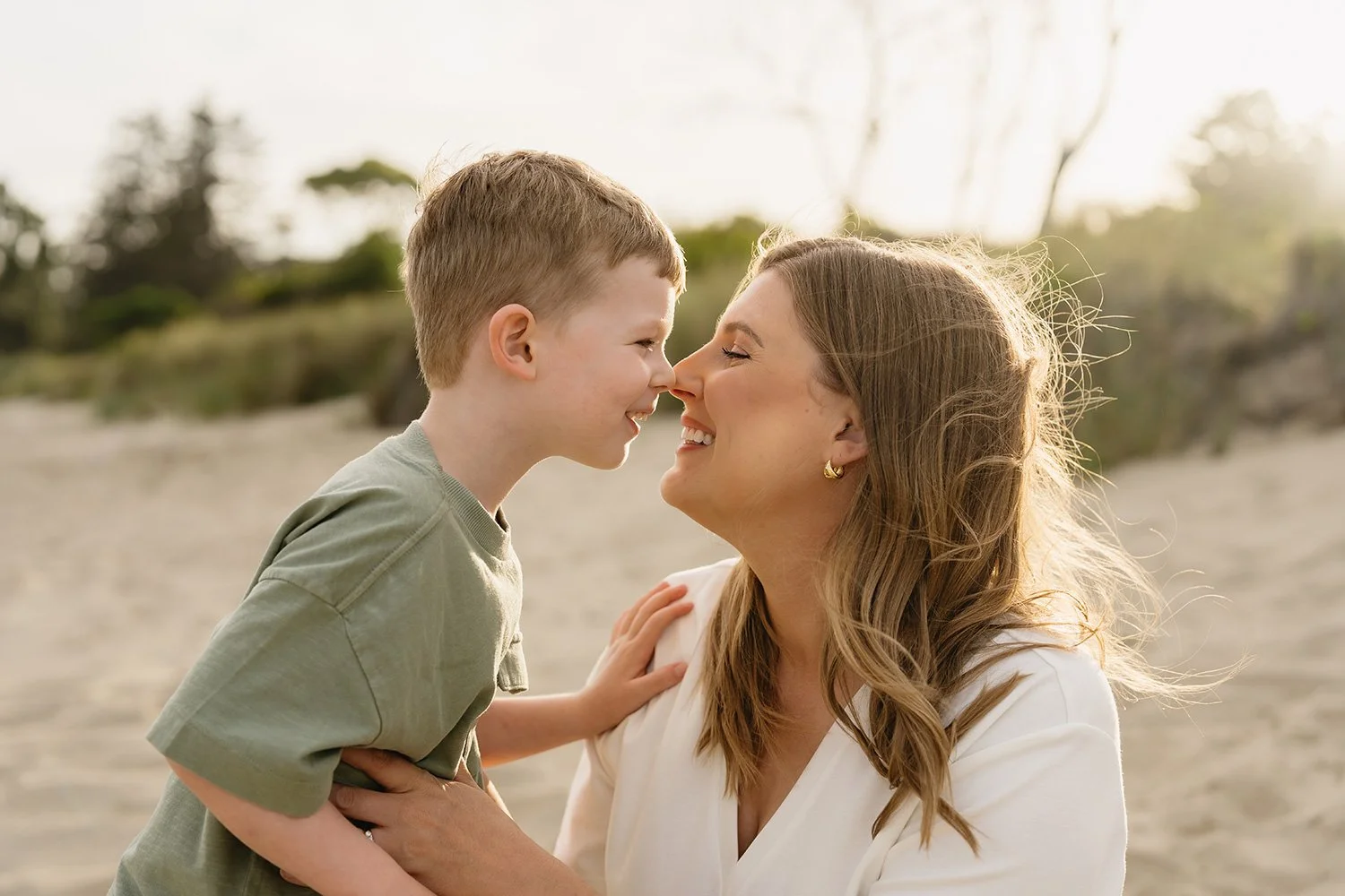 A woman and a young boy with light skin smiling and touching foreheads outdoors on a beach.