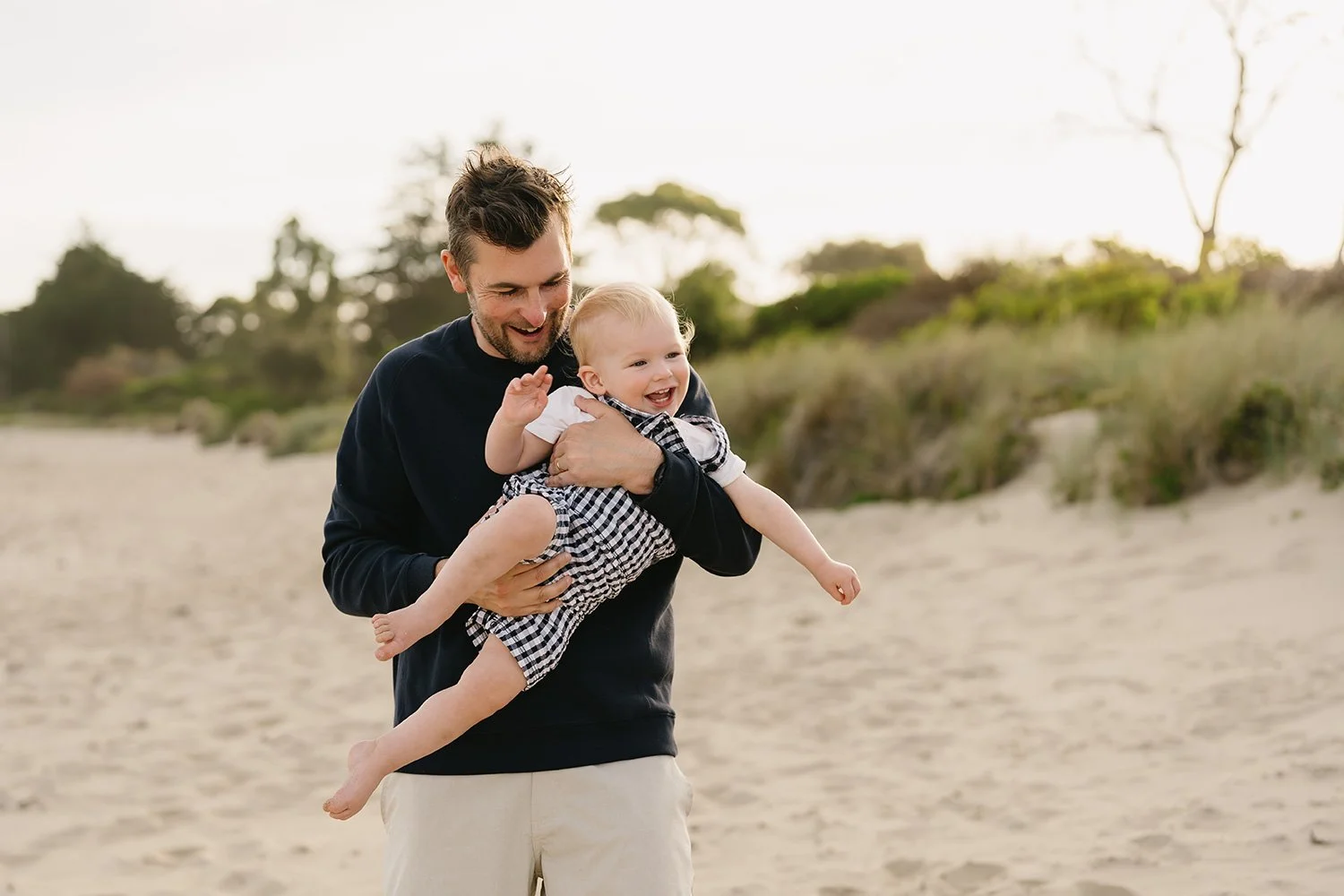 A man holding a laughing baby girl on a sandy beach with trees in the background.