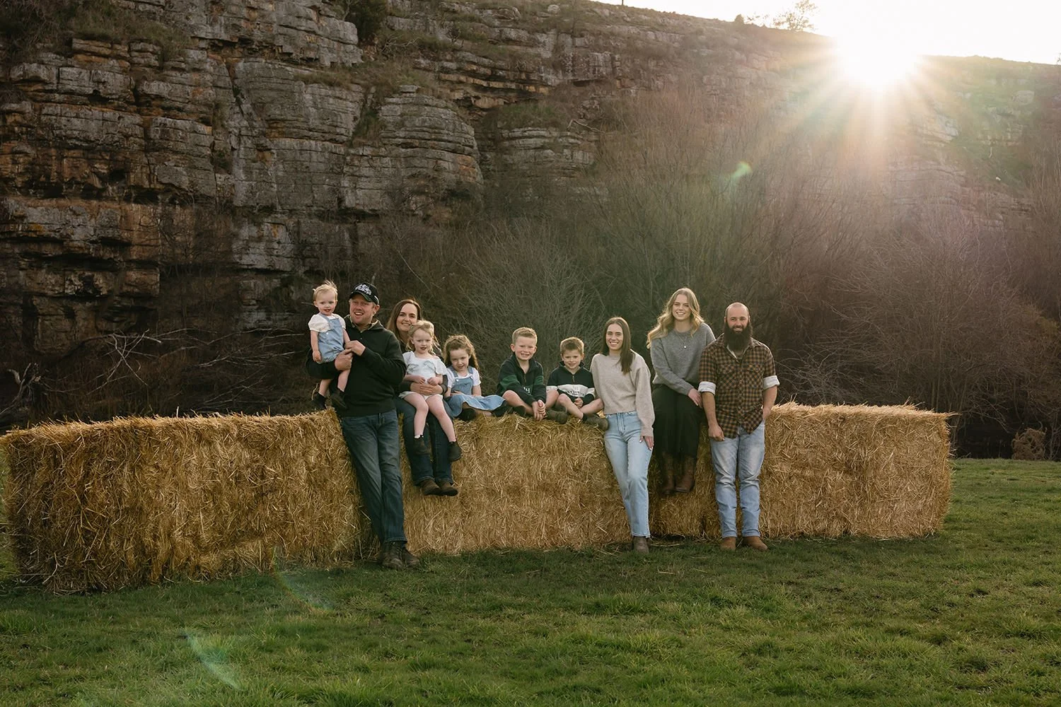 Family of nine people outdoors sitting and standing on a hay bale, with rocky cliffs and leafless trees in the background, during sunset.
