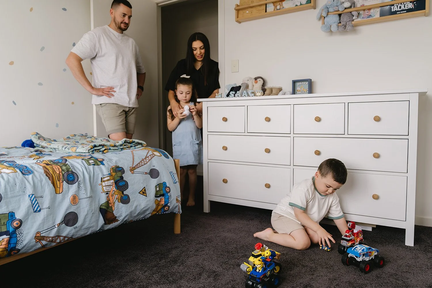 A young boy playing with toy cars on a dark carpeted floor in his bedroom while a man, a woman, and a young girl stand nearby and look on. The room is decorated with a white dresser, a bed with a construction-themed bedspread, and shelves with stuffe