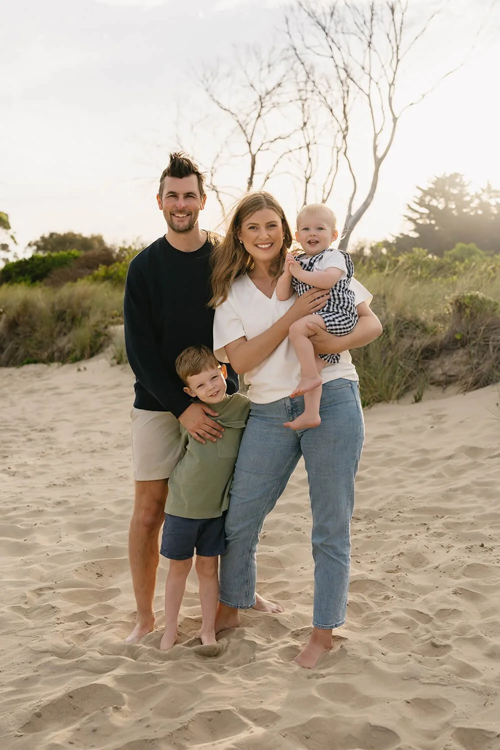 A family of four standing barefoot on a sandy beach with grass and trees in the background during sunset, smiling at the camera.