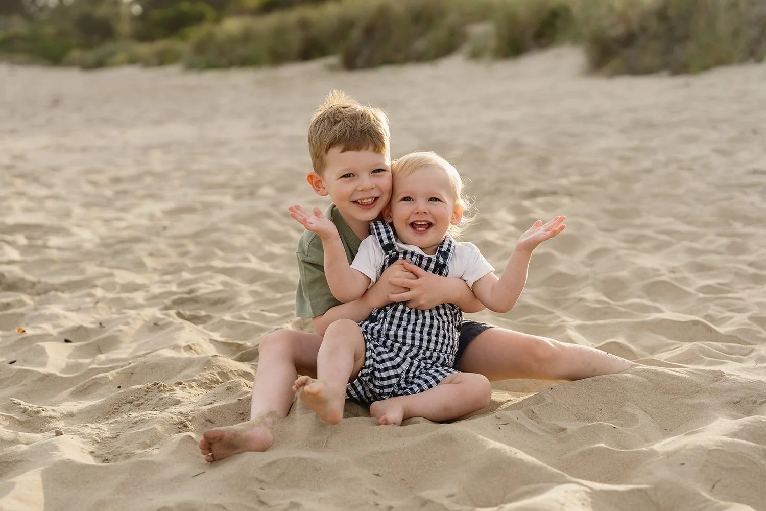 Two young children, a boy and a girl, sit on the sandy beach smiling and playing together, with a background of dunes and grass.