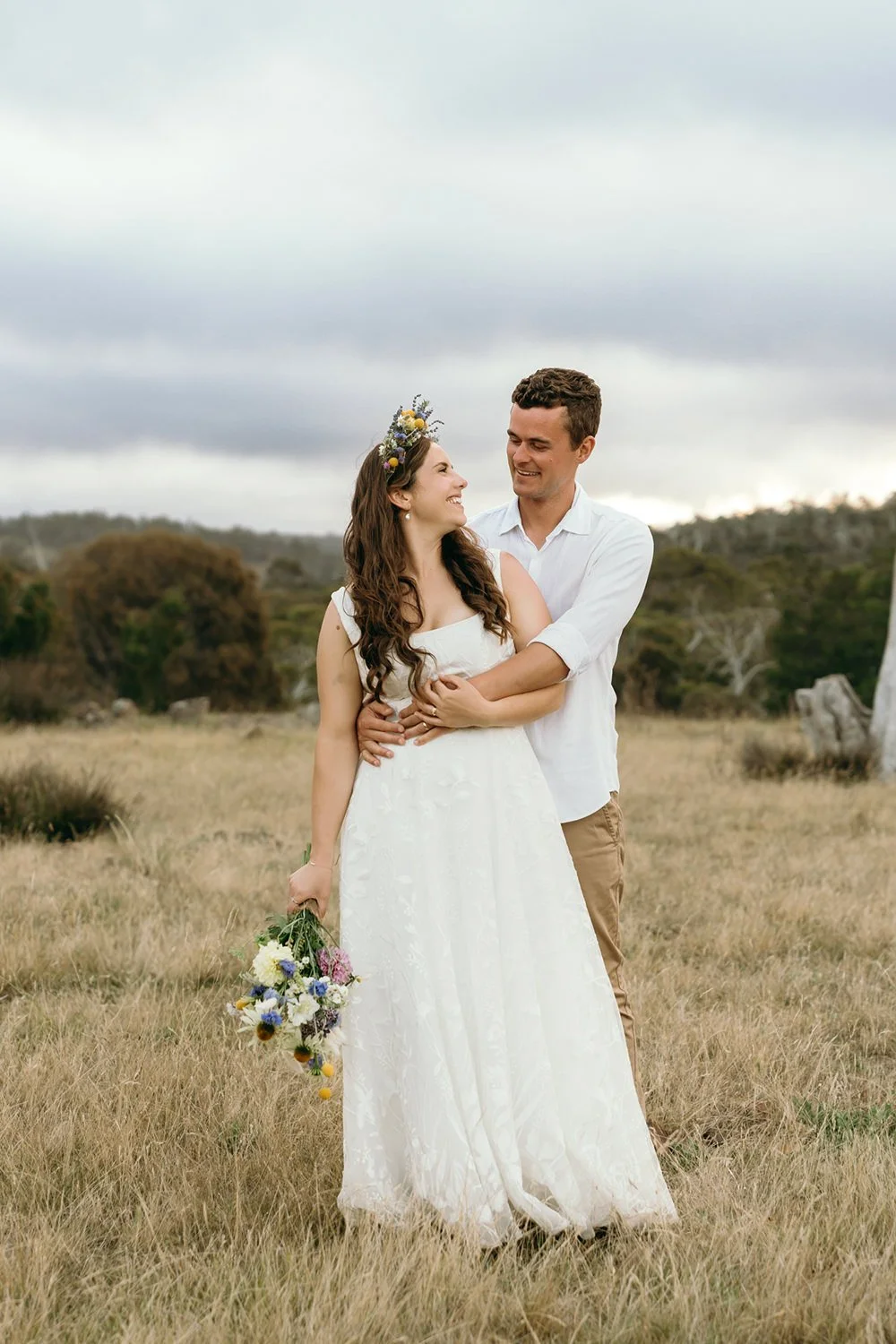 A couple in wedding attire standing in a grassy field, smiling and embracing, with the woman holding a bouquet of flowers and wearing a floral crown, against a cloudy sky and distant trees.