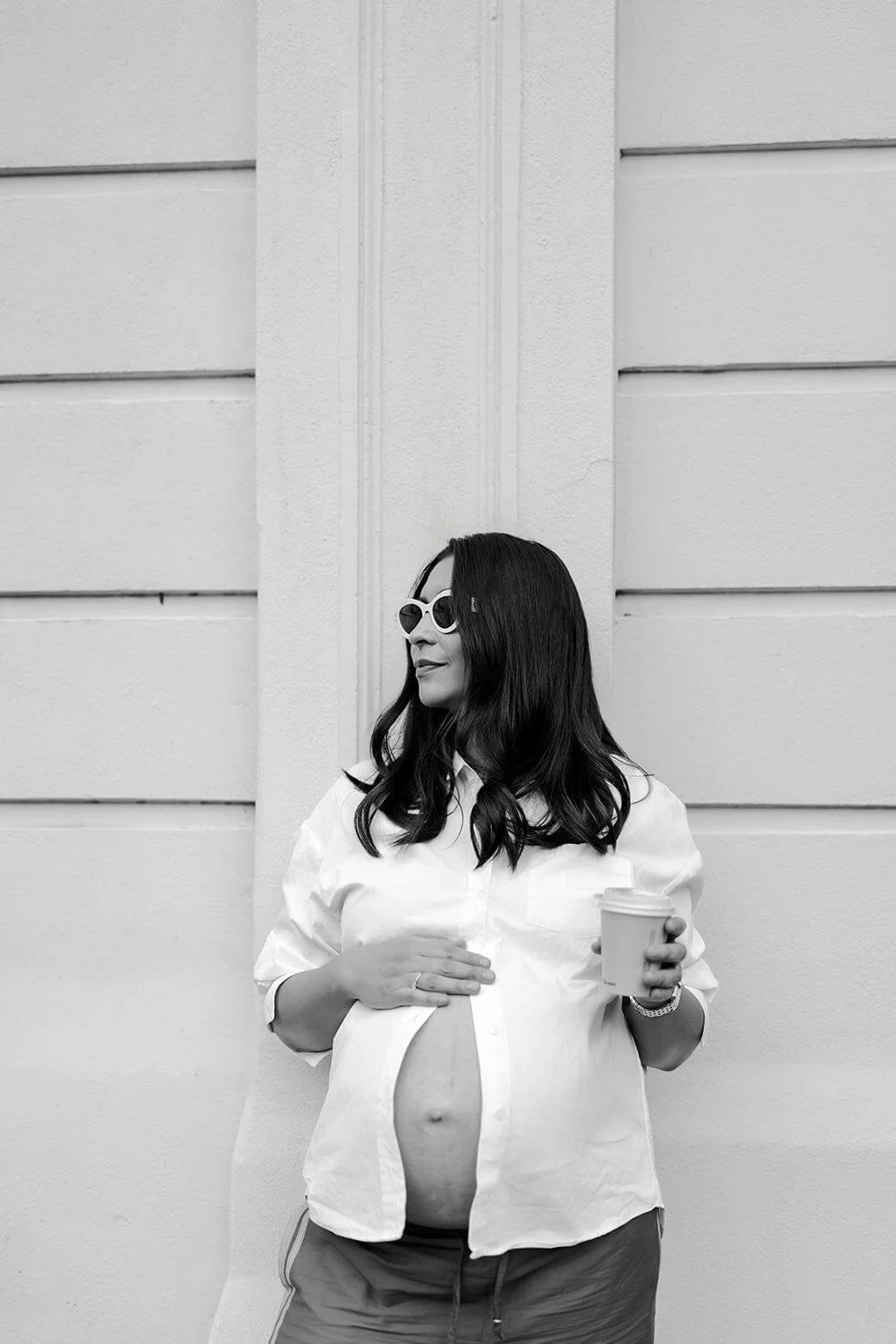 A pregnant woman with long dark hair wearing sunglasses and a white shirt, holding a coffee cup, standing against a textured wall.