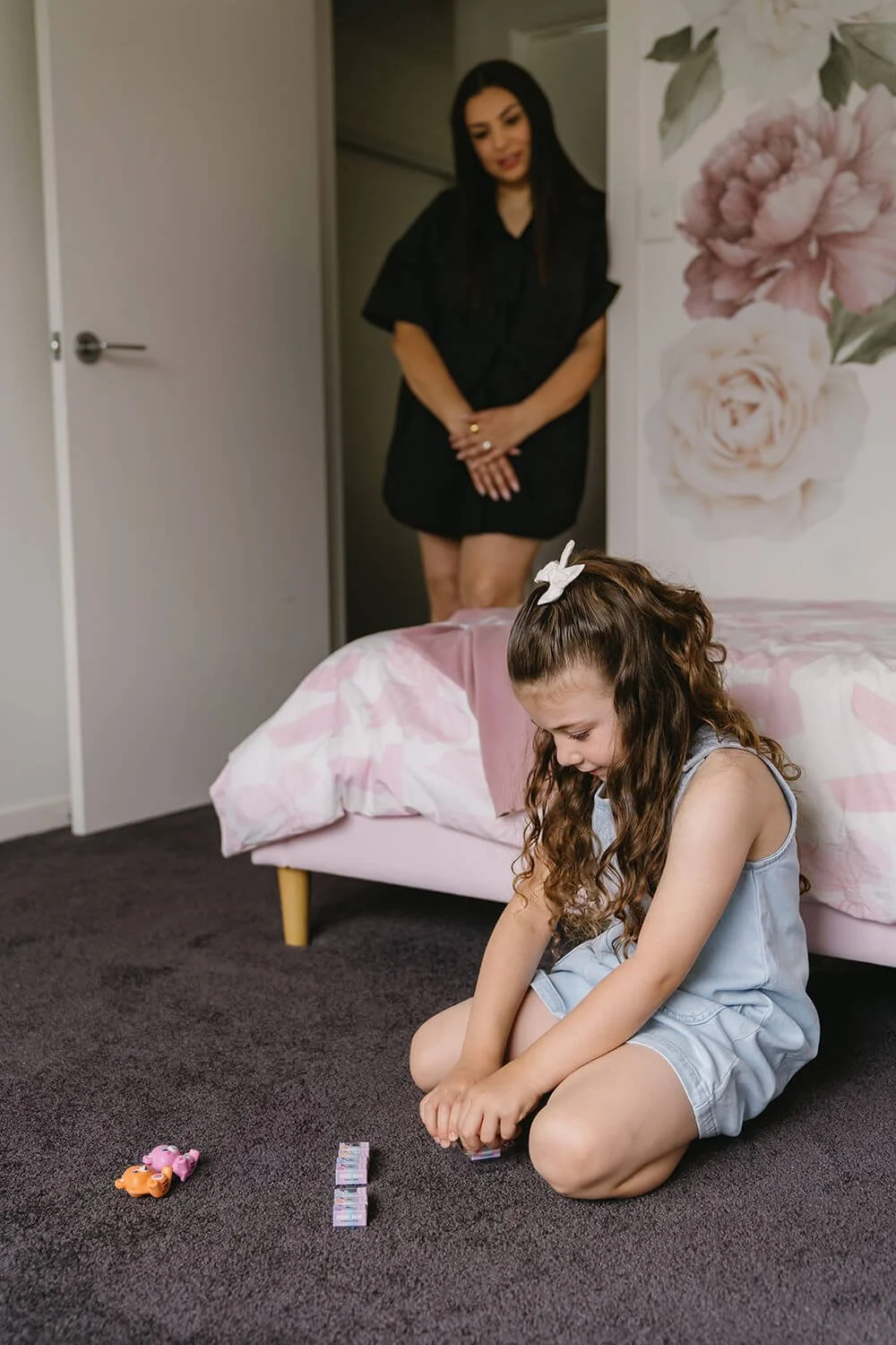 A young girl with curly hair and a white hair bow is sitting on the floor in a bedroom, playing with toys and paper money. An adult woman stands in the background near the door, watching her.